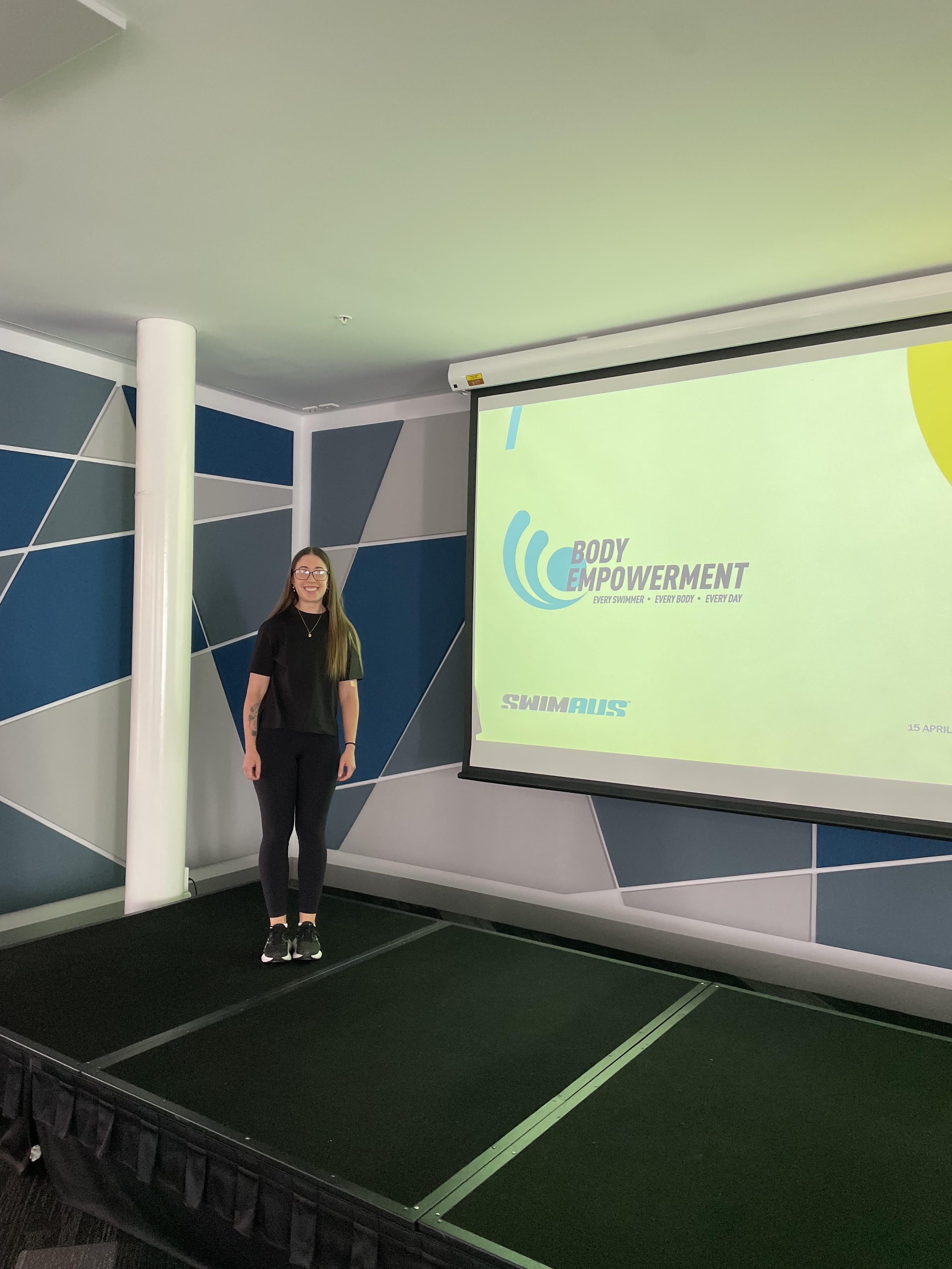 A woman standing on a small stage in front of a presentation screen that displays a logo and text about Body Empowerment and swimming.