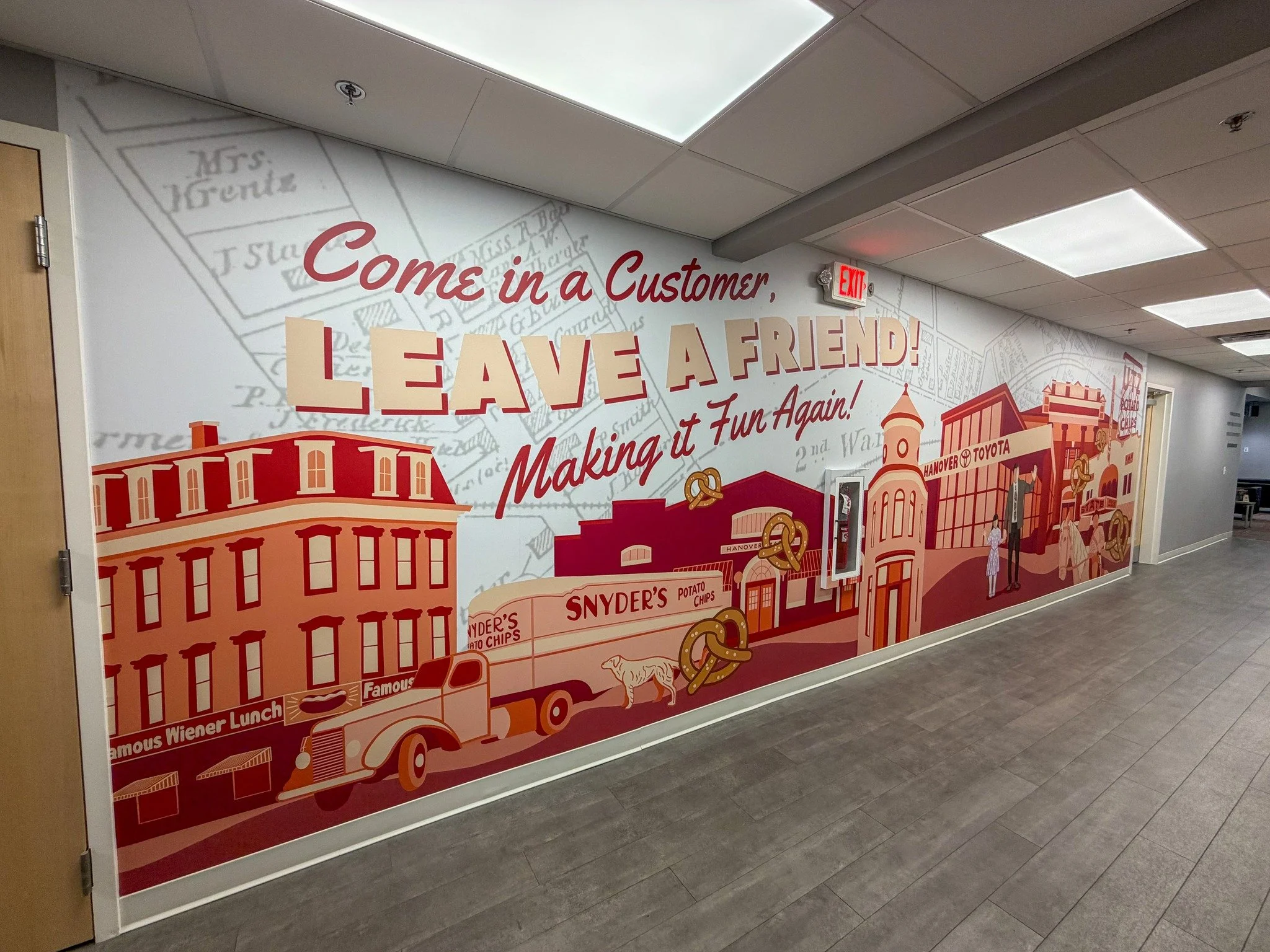 Decorative wall mural with red and beige graphics and text that says 'Come in a Customer, Leave a Friend! Making it Fun Again!' featuring illustrations of buildings, a truck, a dog, and people in a town setting. Hanover Toyota in Hanover, PA.