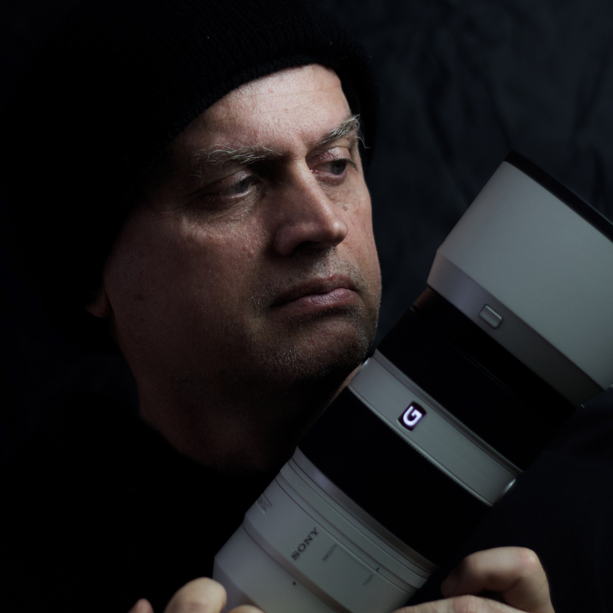 A man with a serious expression is lying on a dark surface, holding a large professional camera with a telephoto lens close to his face.