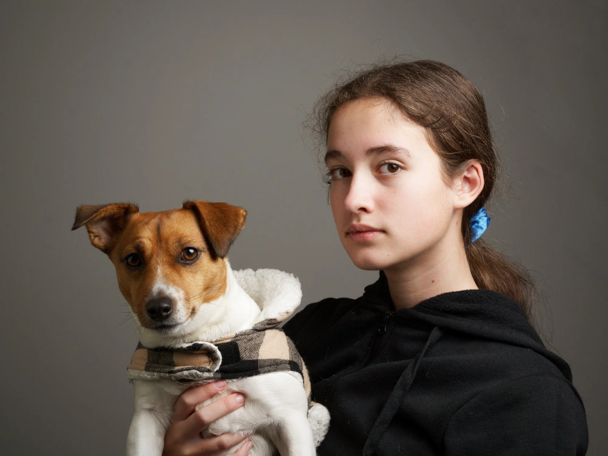 A young woman with brown hair tied back with a blue hair tie, wearing a black hoodie, holding a small brown and white dog wearing a plaid coat, against a plain gray background.