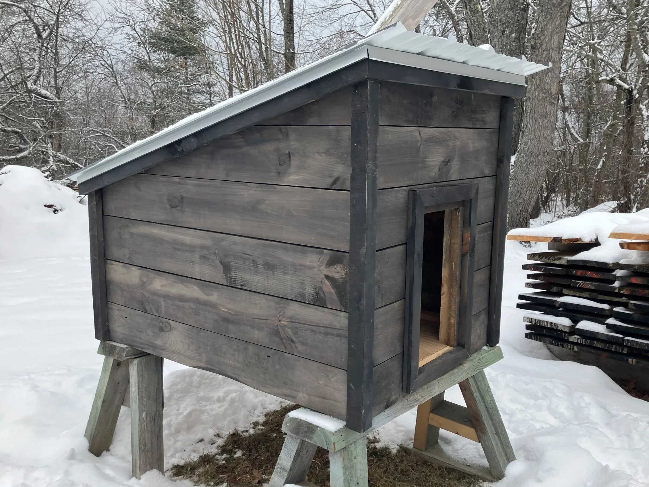 A wooden doghouse with a metal roof, set outdoors in a snowy area, with stacked wood nearby and leafless trees in the background.