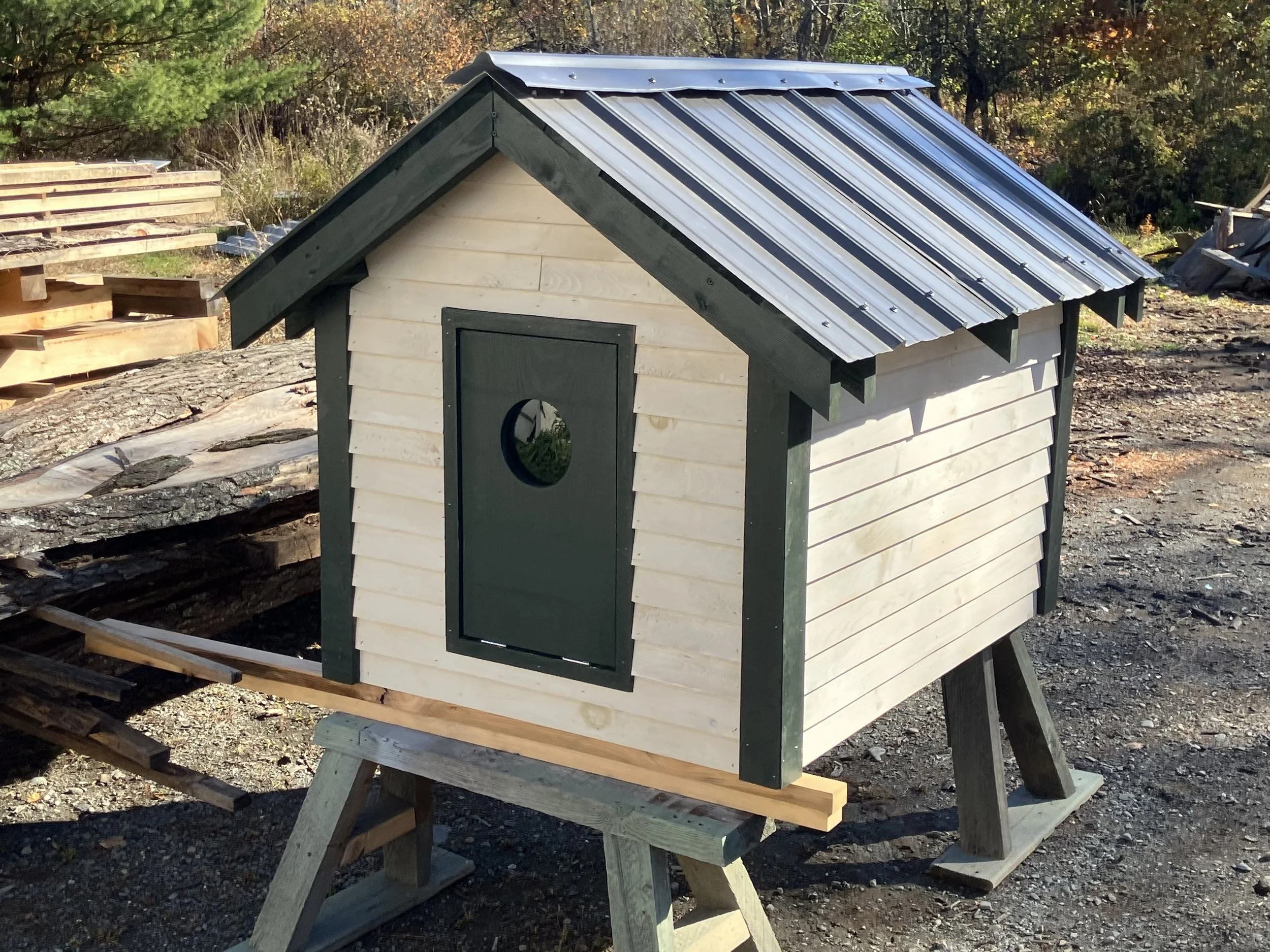 A small wooden pet house with a sloped metal roof on a work table outdoors, with wood logs and trees in the background.