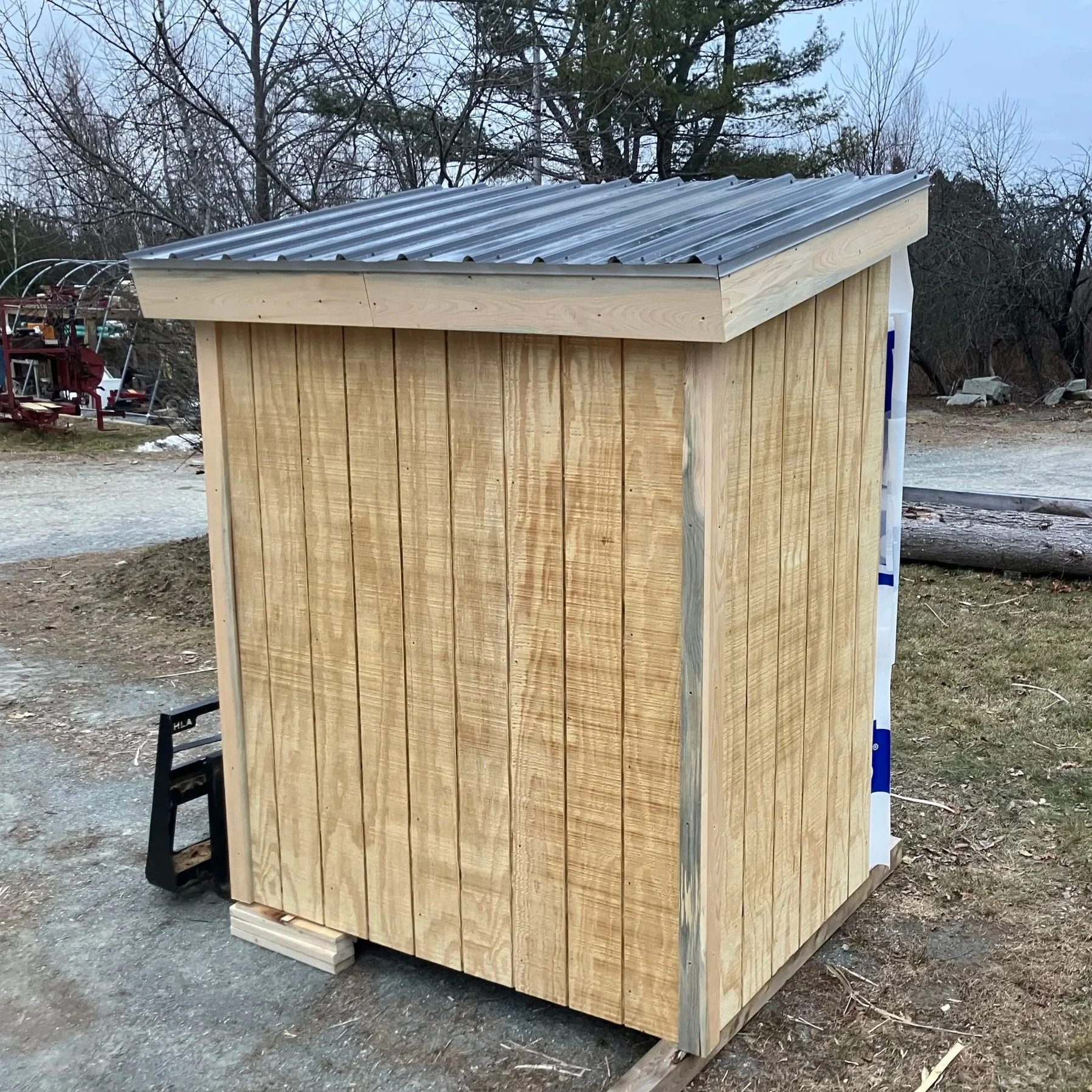 Unpainted wooden outdoor storage shed with a metal roof, situated on a patch of dirt and grass outside in a rural area.