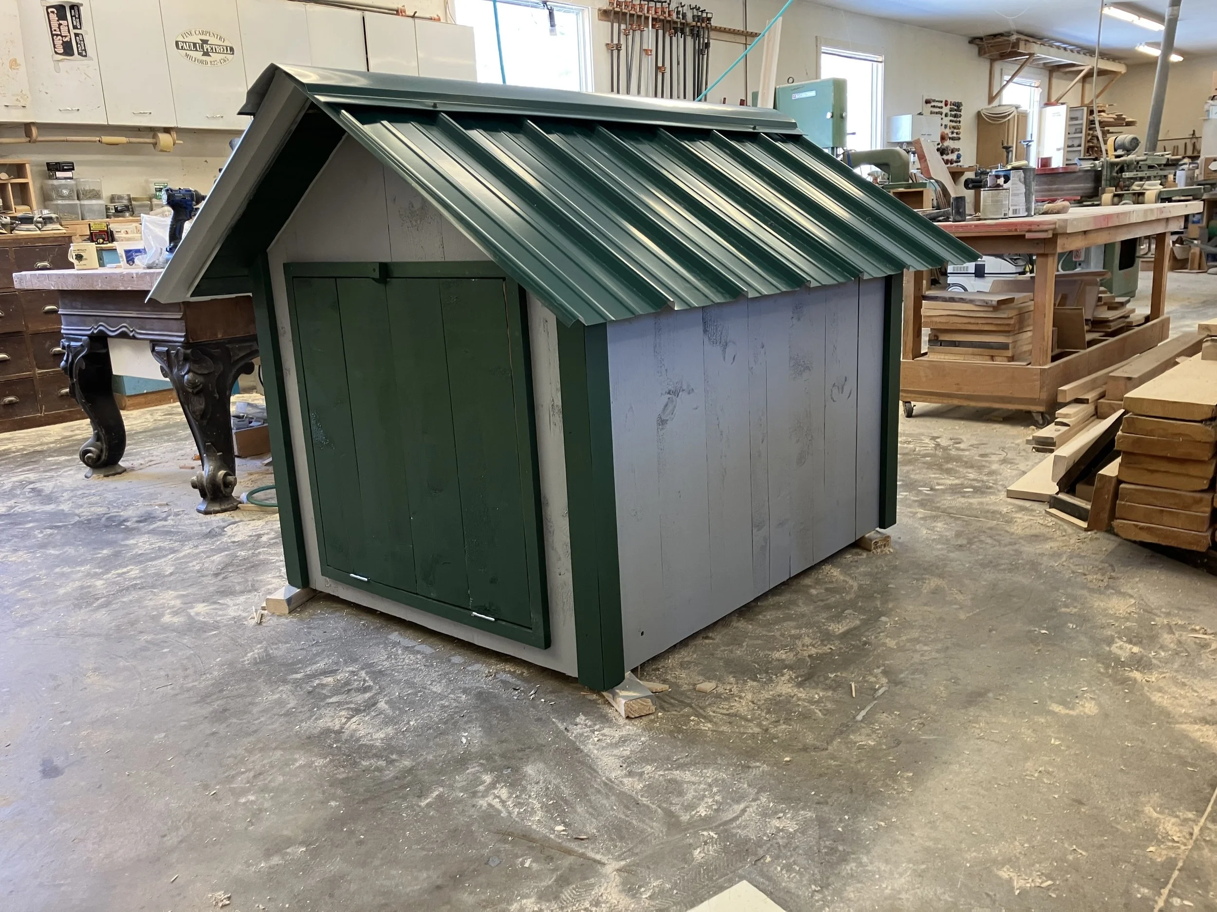 A small green and white playhouse with a sloped green metal roof is situated in a wood workshop with various tools and wooden materials in the background.