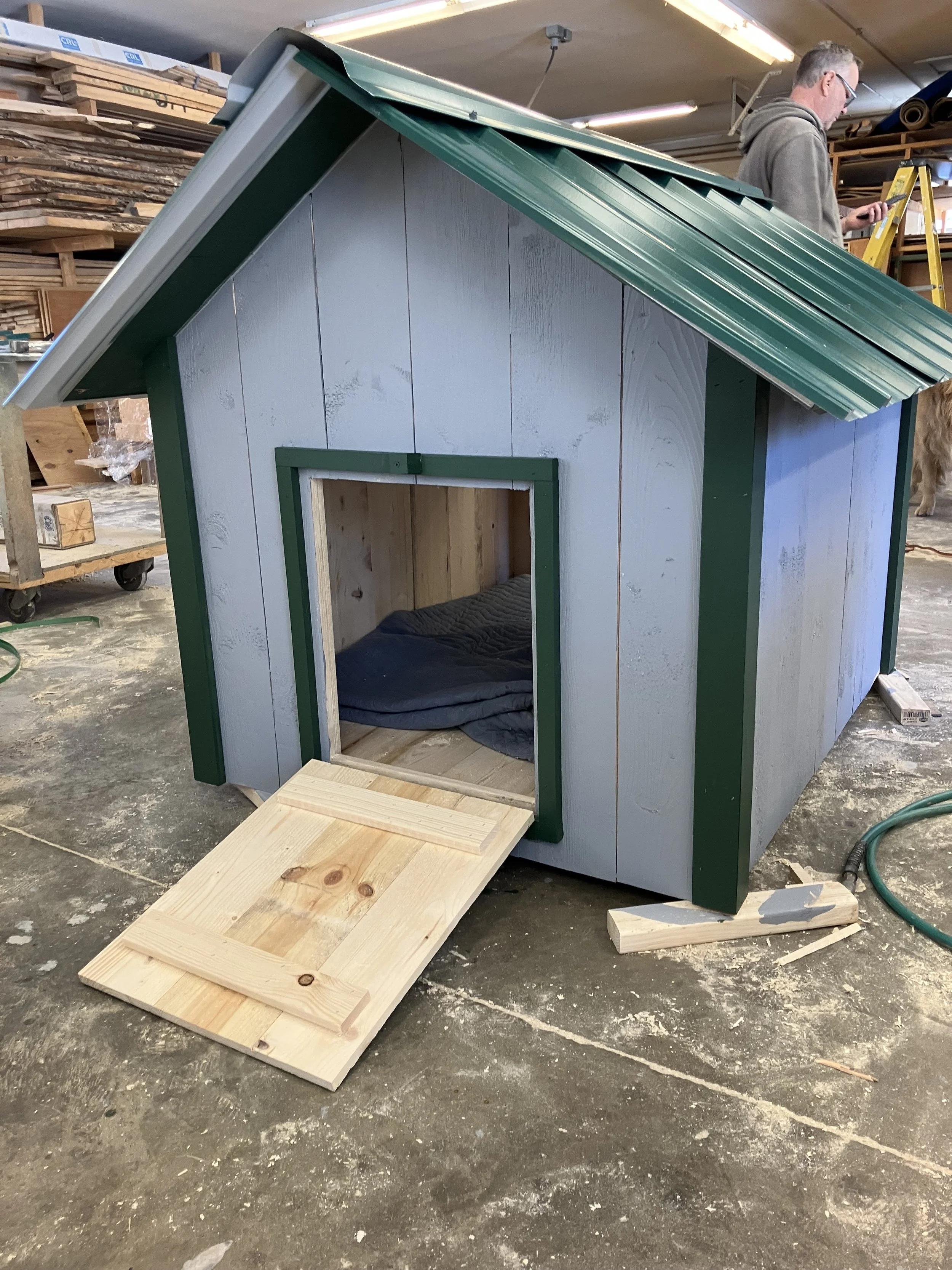 A small dog house with a green trim and a ramp entrance in a woodworking workshop. A man in a gray hoodie stands to the right, working with a yellow ladder. The workshop is filled with wood and tools.