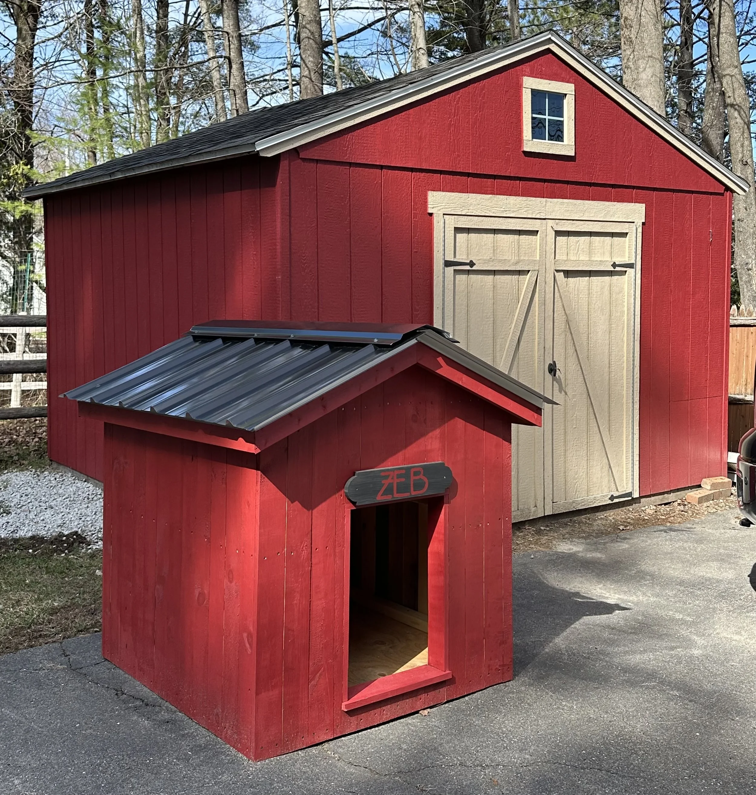 A red wooden shed with a small window near the roof and a double-door entrance, located outdoors in a wooded area with trees and a partly cloudy sky in the background.