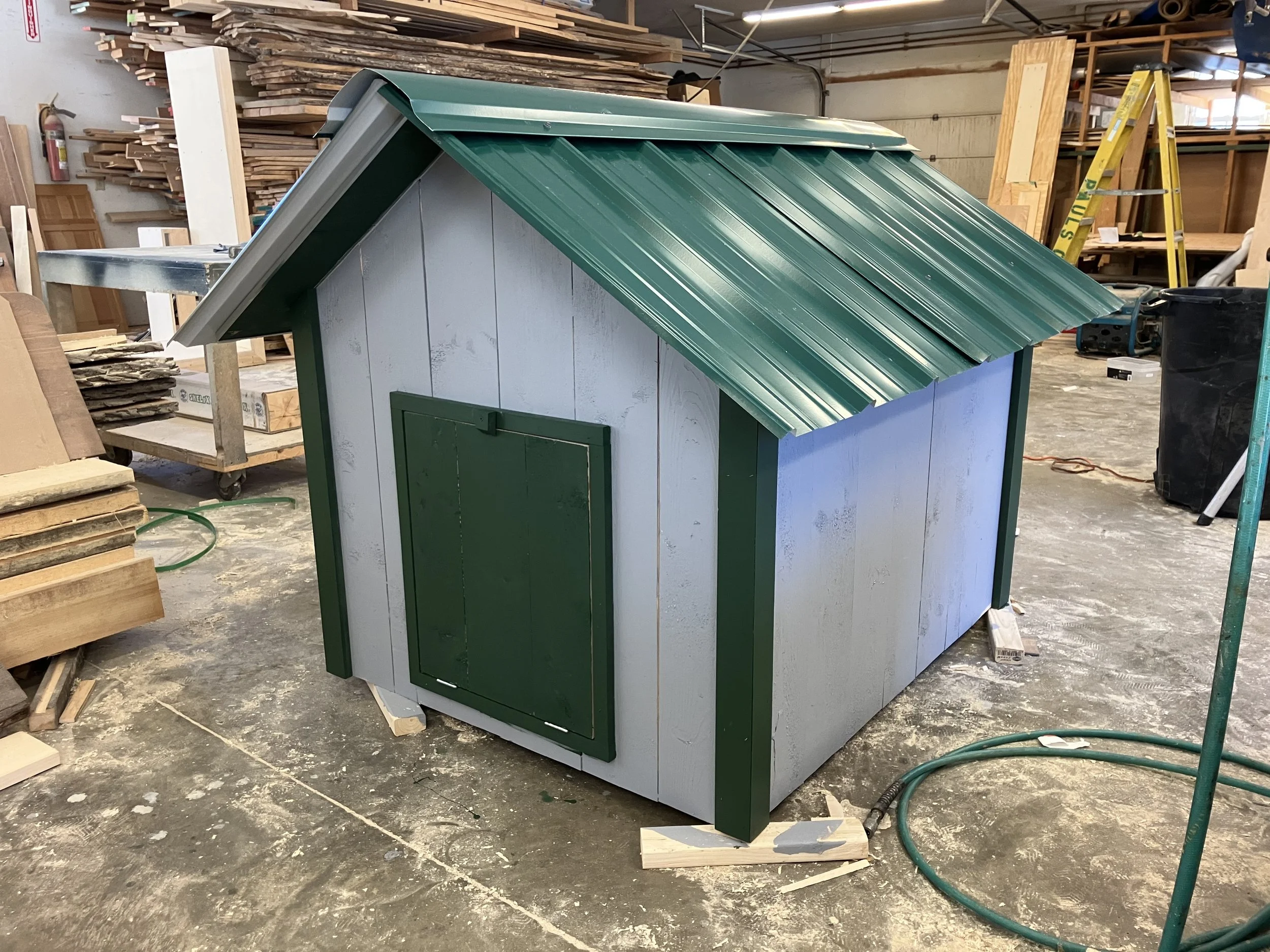 A small house with a green metal roof and white walls, situated in a woodworking workshop with stacks of wood and tools in the background.