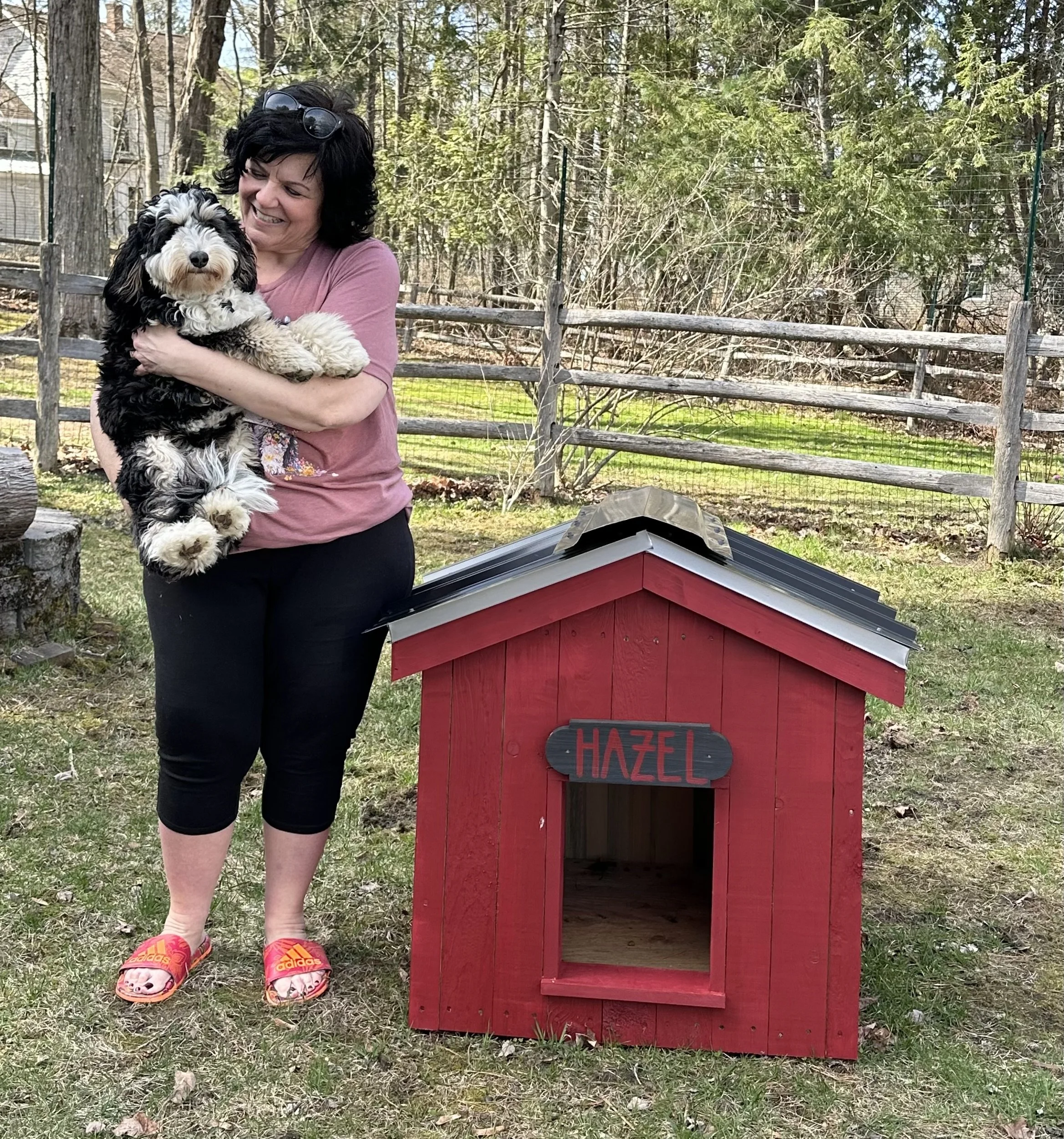Woman holding a black and white fluffy dog outdoors near a red doghouse with a black sign that says "Hazel." She is smiling and wearing a pink shirt, black pants, colorful sandals, and sunglasses on her head. The background shows trees, a wooden fence, and a house.