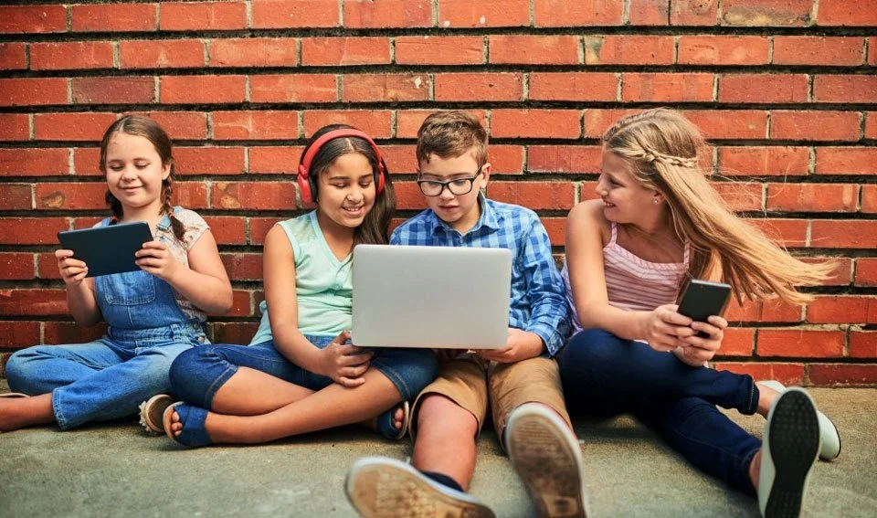 Kids sitting near brick wall streaming with laptop, tablet, and phone