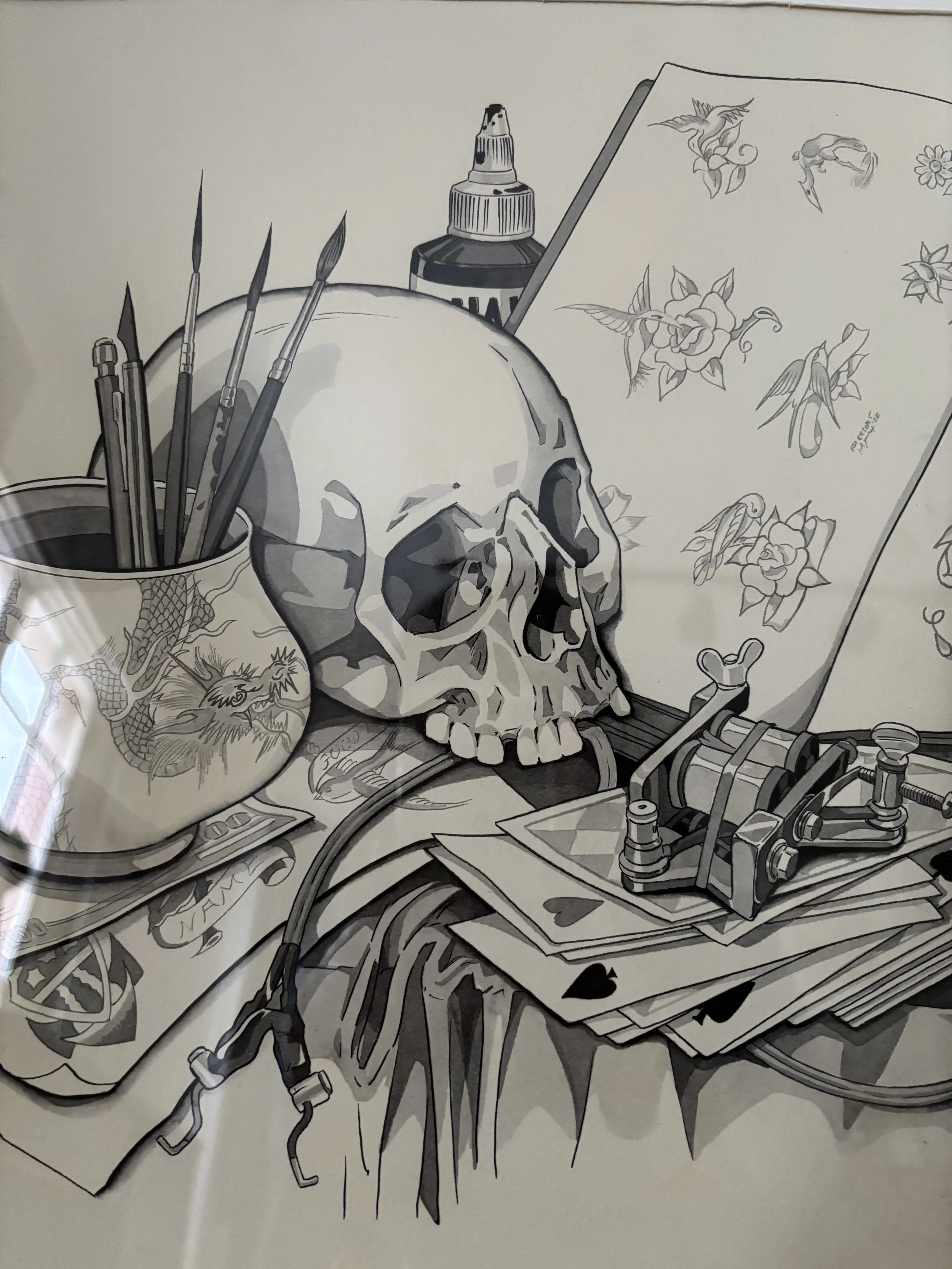 Black and white drawing of an artist's workspace including a human skull, paintbrushes in a jar, tattoo designs featuring roses and hummingbirds, and tattoo equipment on a table.