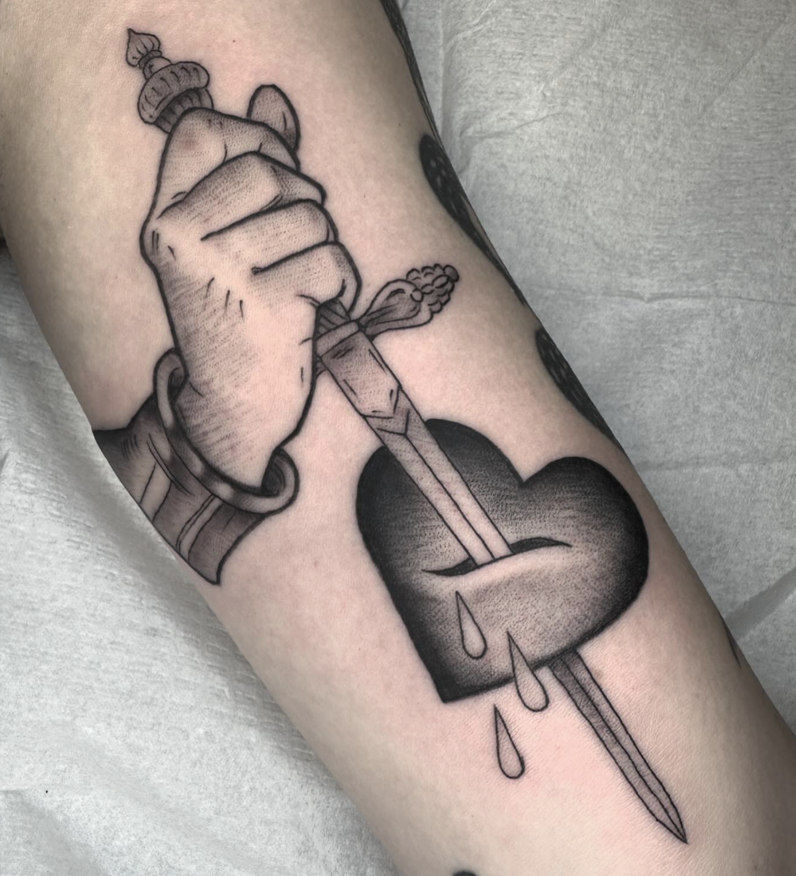 A tattoo of a hand holding a dagger piercing a heart with tears falling from it.