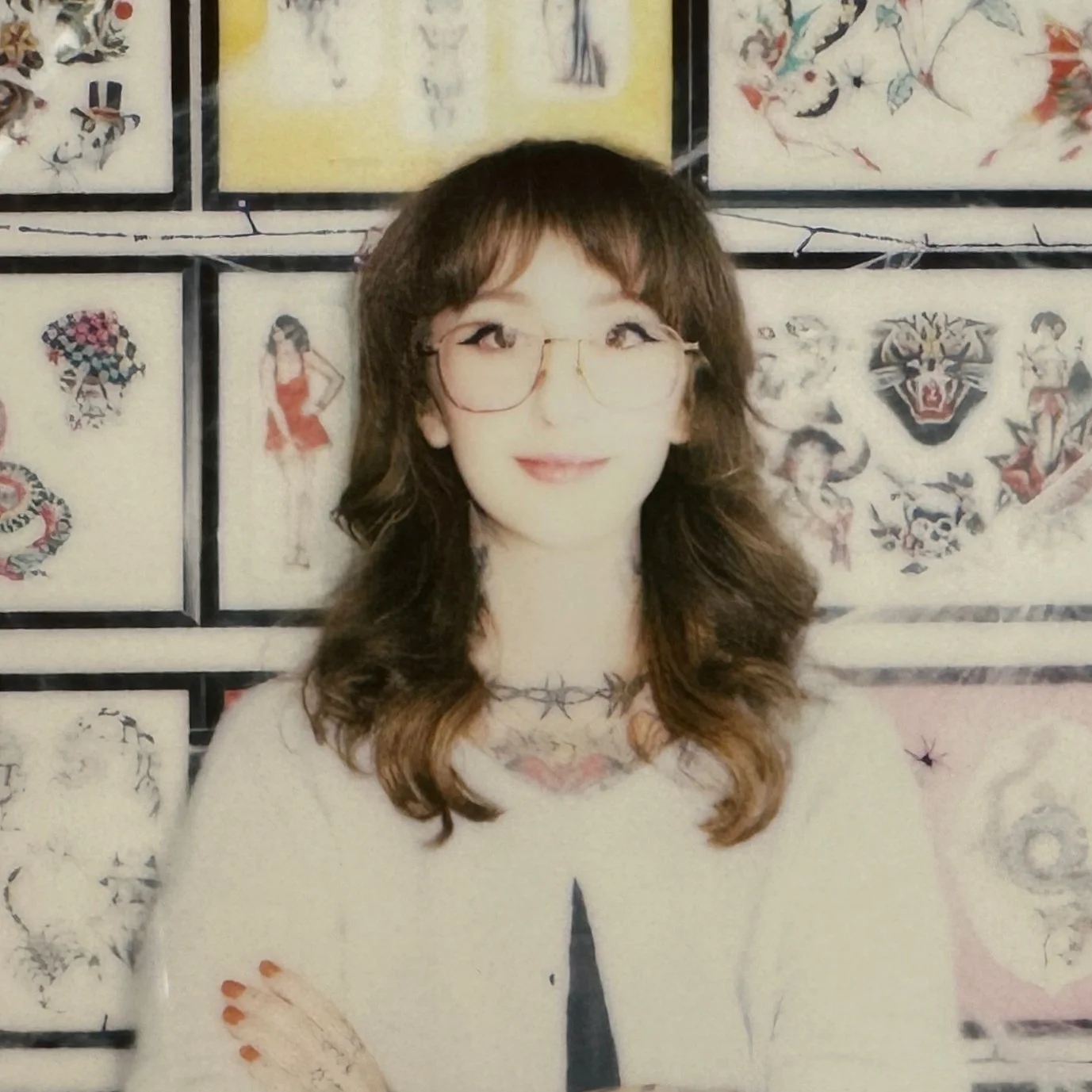 A woman with brown hair, glasses, and a tattoo necklace, smiling in front of a wall with framed traditional tattoo flash art.