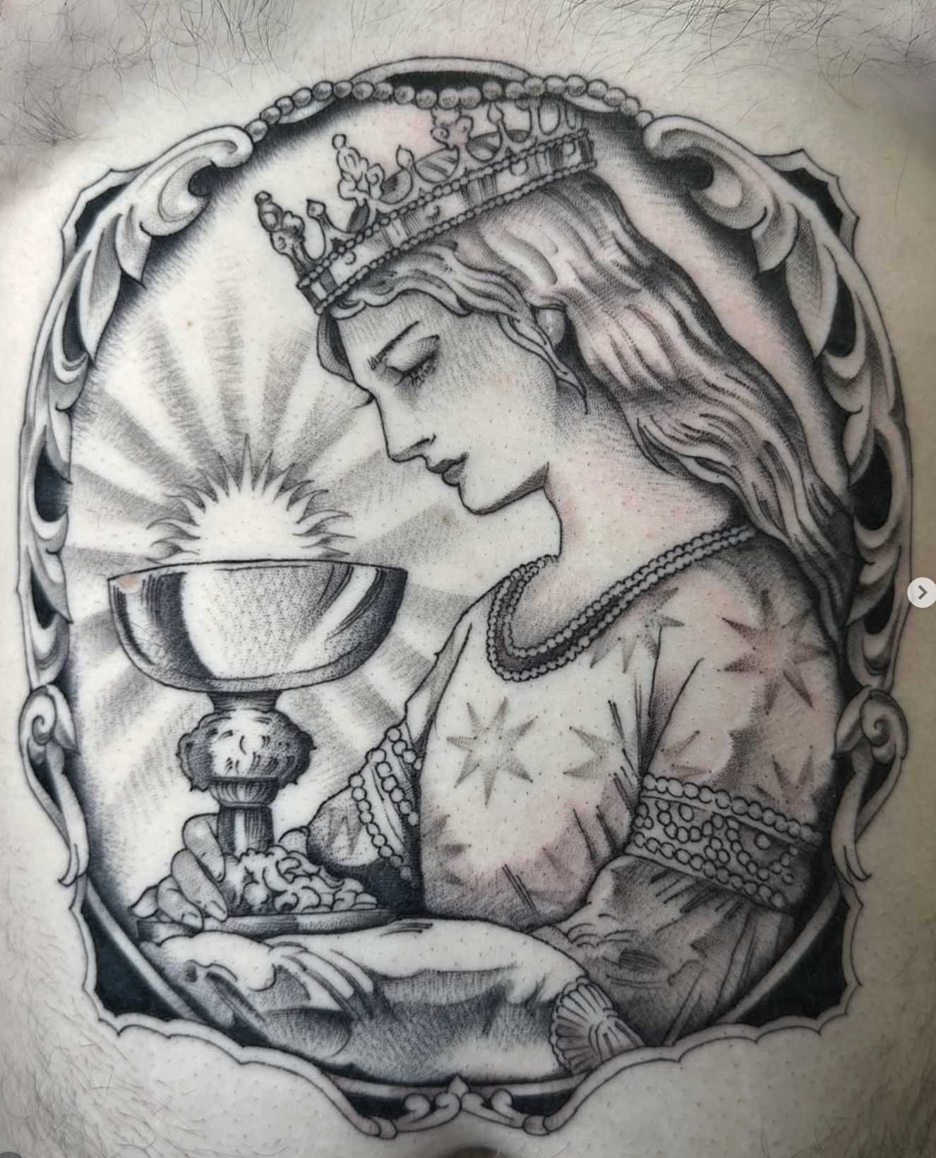 Tattoo of a woman wearing a crown, holding a chalice with a radiant light emerging from it, surrounded by ornate decorative borders.