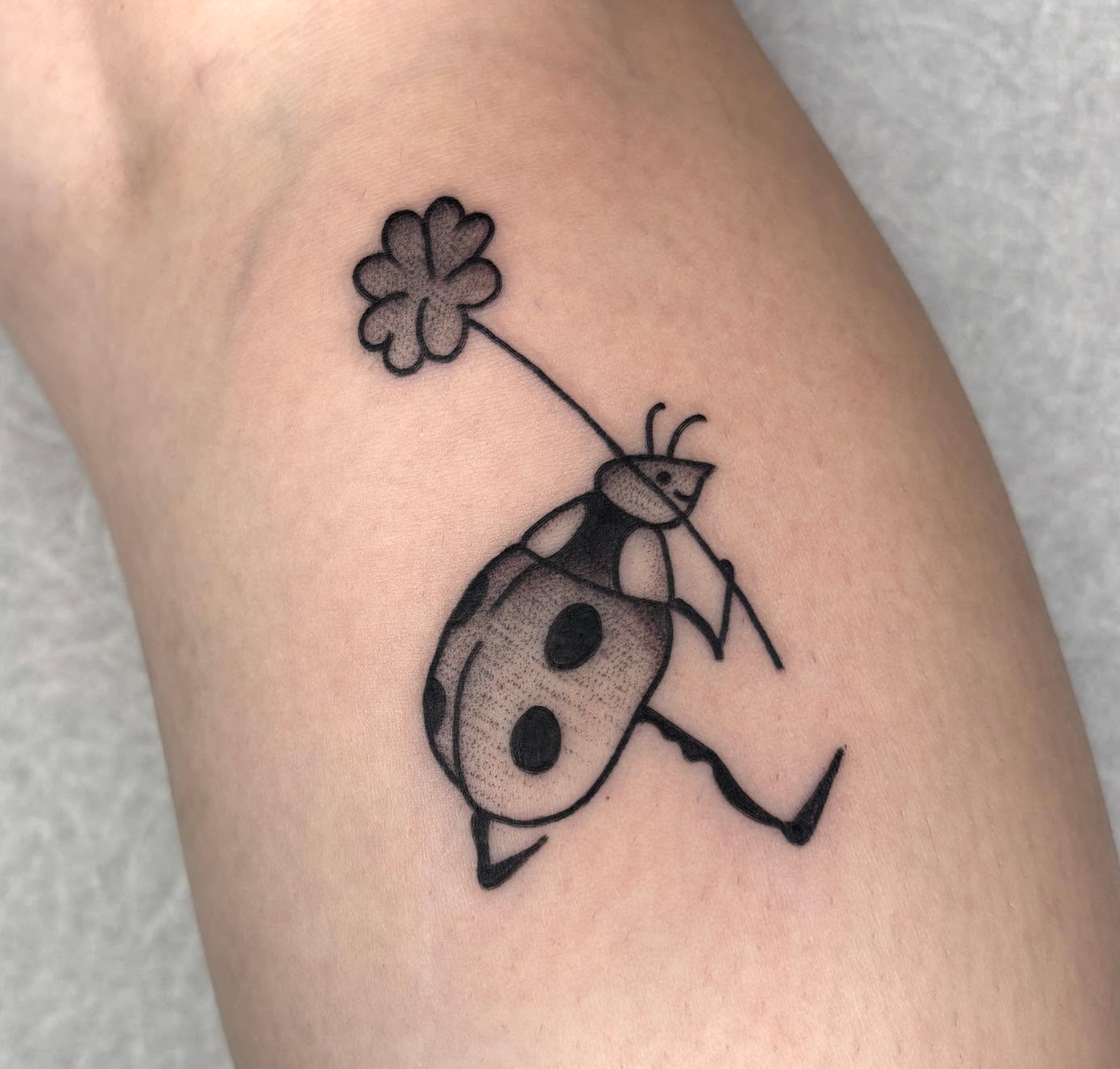 Tattoo of a ladybug with a four-leaf clover on a stem in its mouth, featuring simple black outline and minimal shading on skin.
