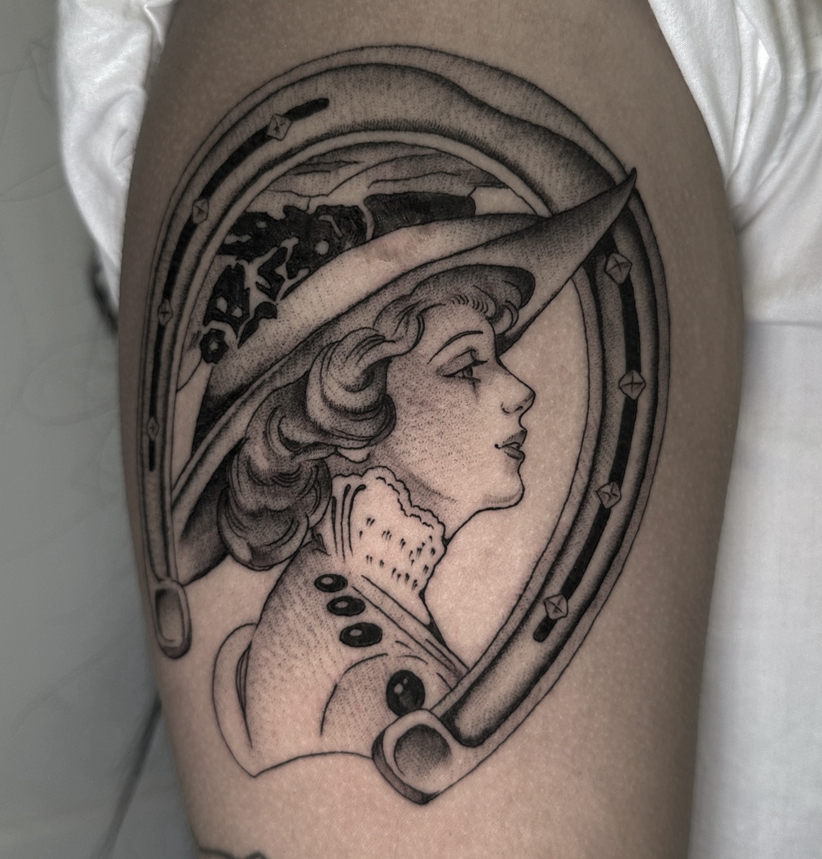 Black and gray tattoo of a woman with wavy hair wearing a large witch hat, in profile view, surrounded by a decorative oval frame.