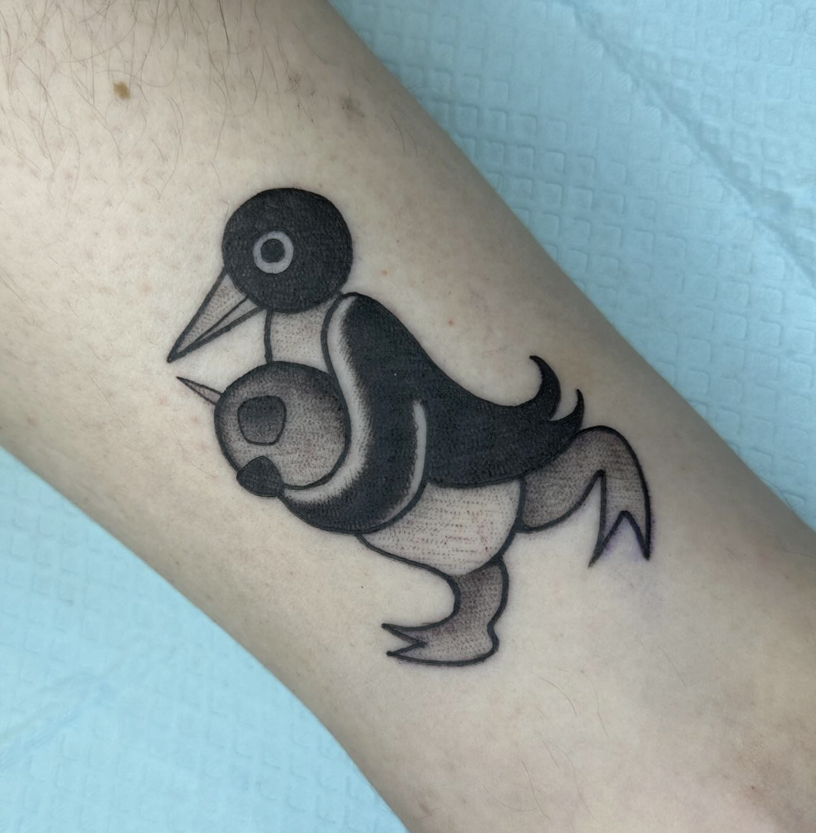 A tattoo of a cartoonish fish with a bird's head, large eye, and a beak, featuring a black and gray color scheme.