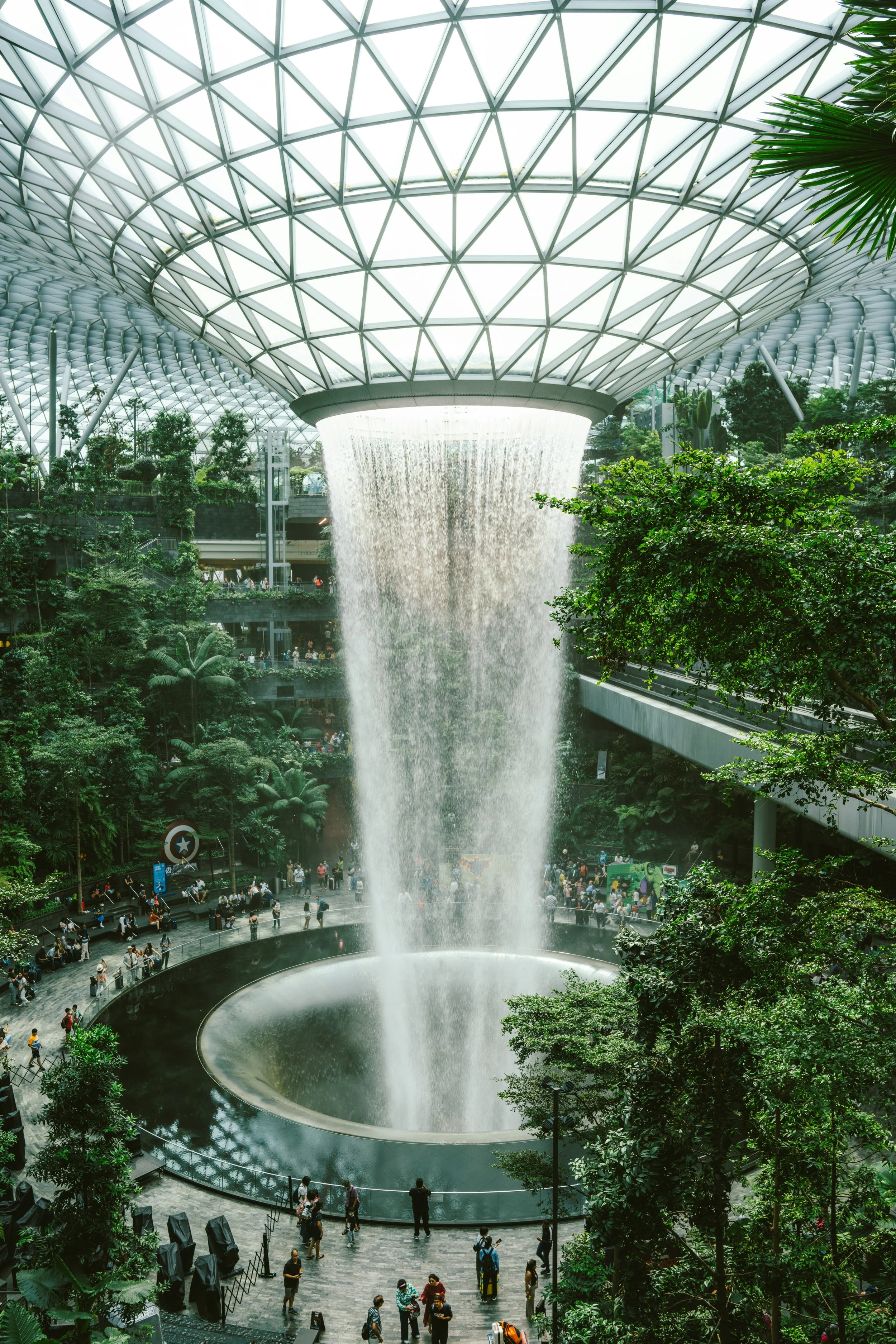Indoor waterfall with a dome glass ceiling, surrounded by lush greenery and visitors inside a large atrium.