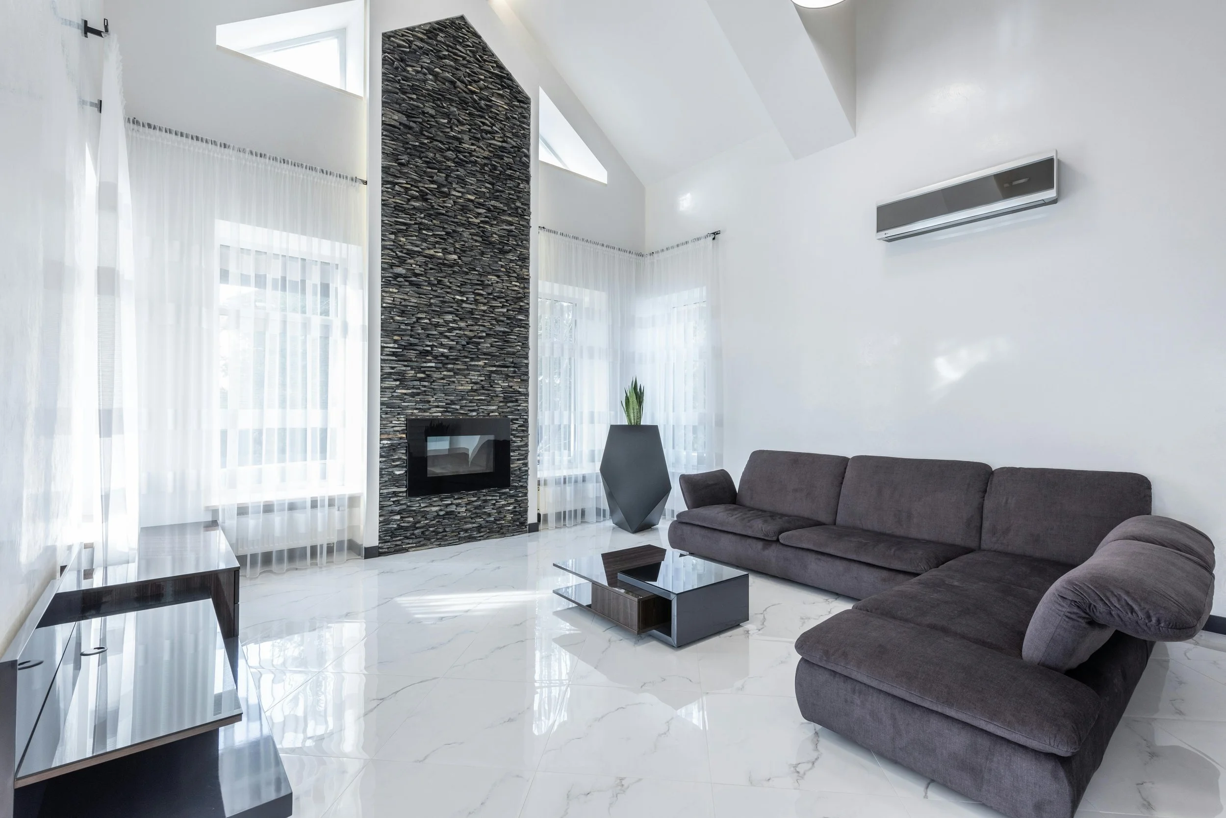 Modern living room with white walls, large windows with sheer curtains, a dark gray sectional sofa, a black and glass coffee table, and a decorative stone fireplace with a built-in firebox. There is a tall black vase with a green plant and a wall-mounted air conditioning unit.