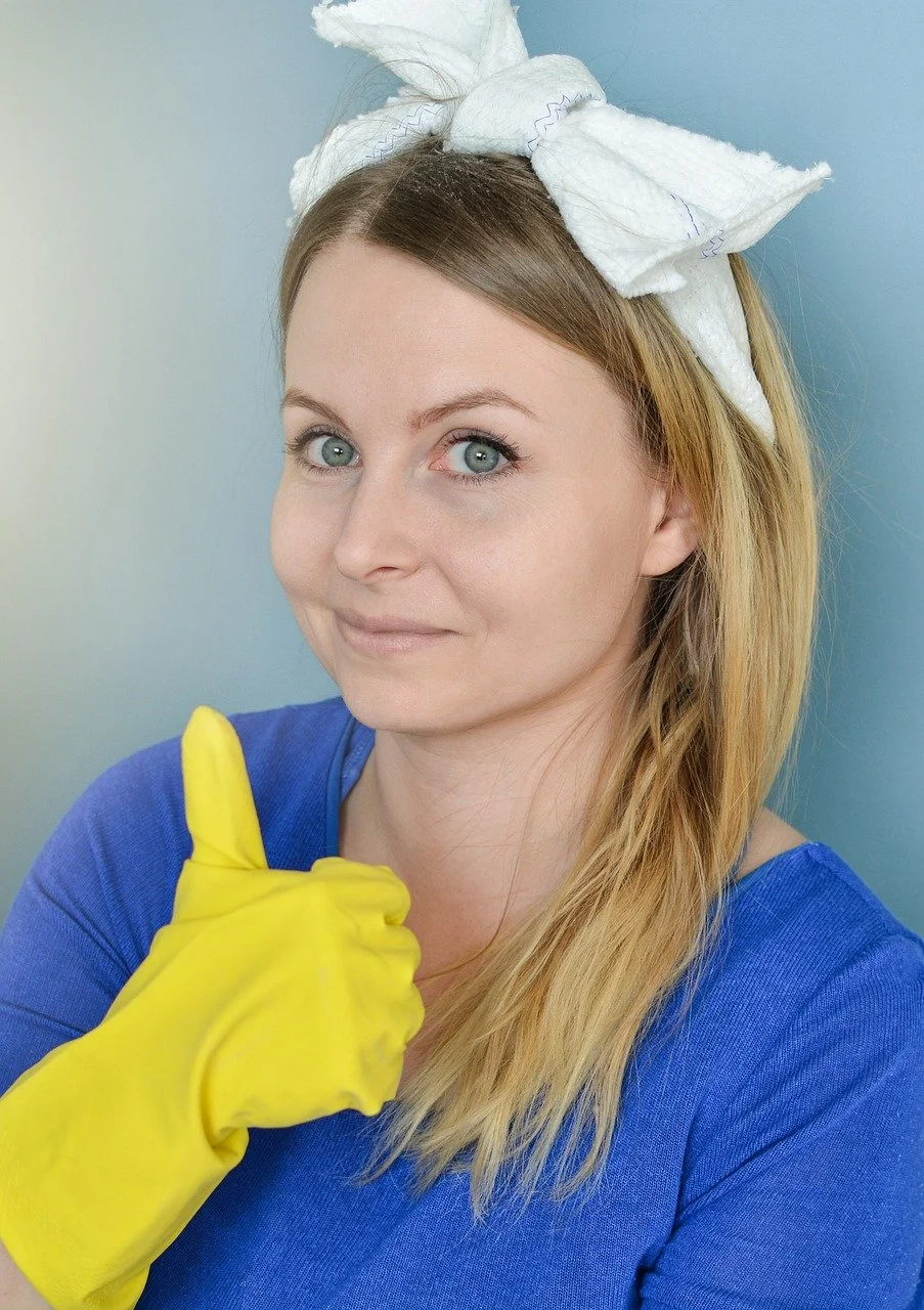 A woman with blonde hair wearing a white headband and a blue shirt, giving a thumbs-up and wearing yellow gloves against a blue background.