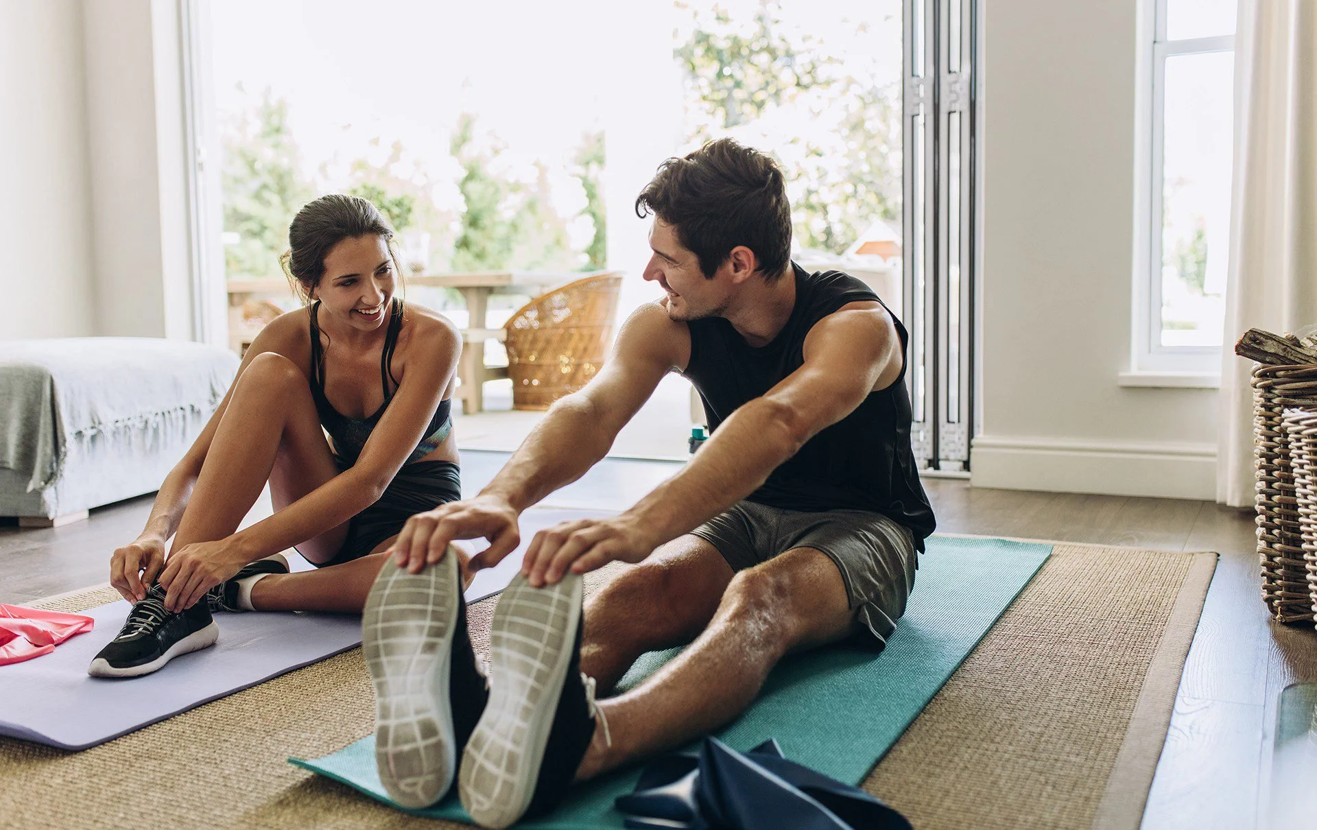 A man and woman sitting on exercise mats stretching indoors by a large open door, smiling and engaging with each other as they stretch their legs.