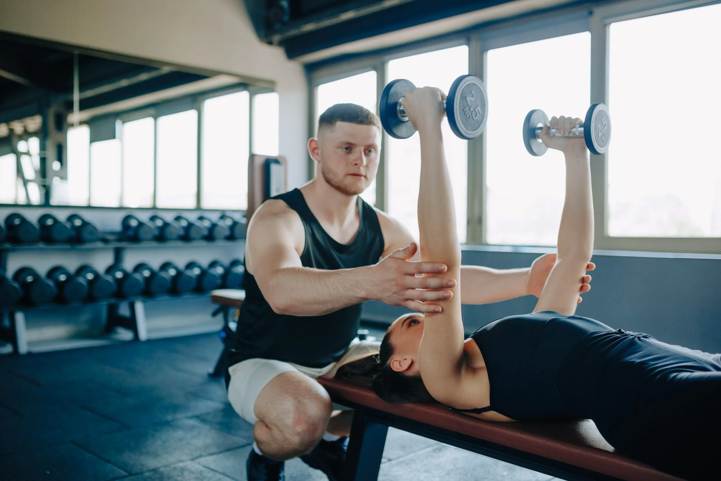 Man helping woman with shoulder exercises using dumbbells in gym.