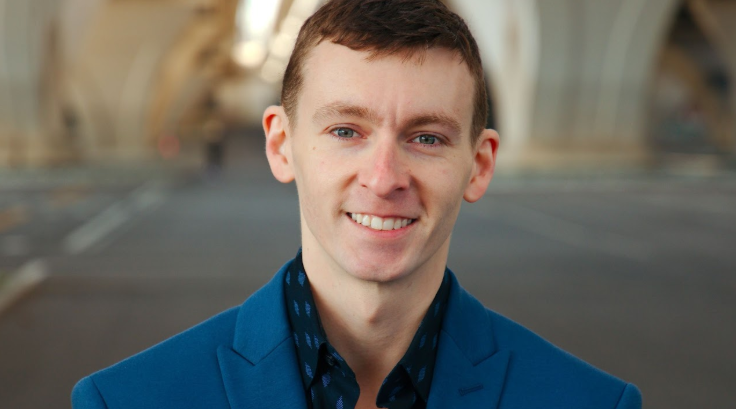 A young man with short brown hair, fair skin, and blue eyes, smiling and wearing a blue blazer, standing outdoors on a street with blurred background.