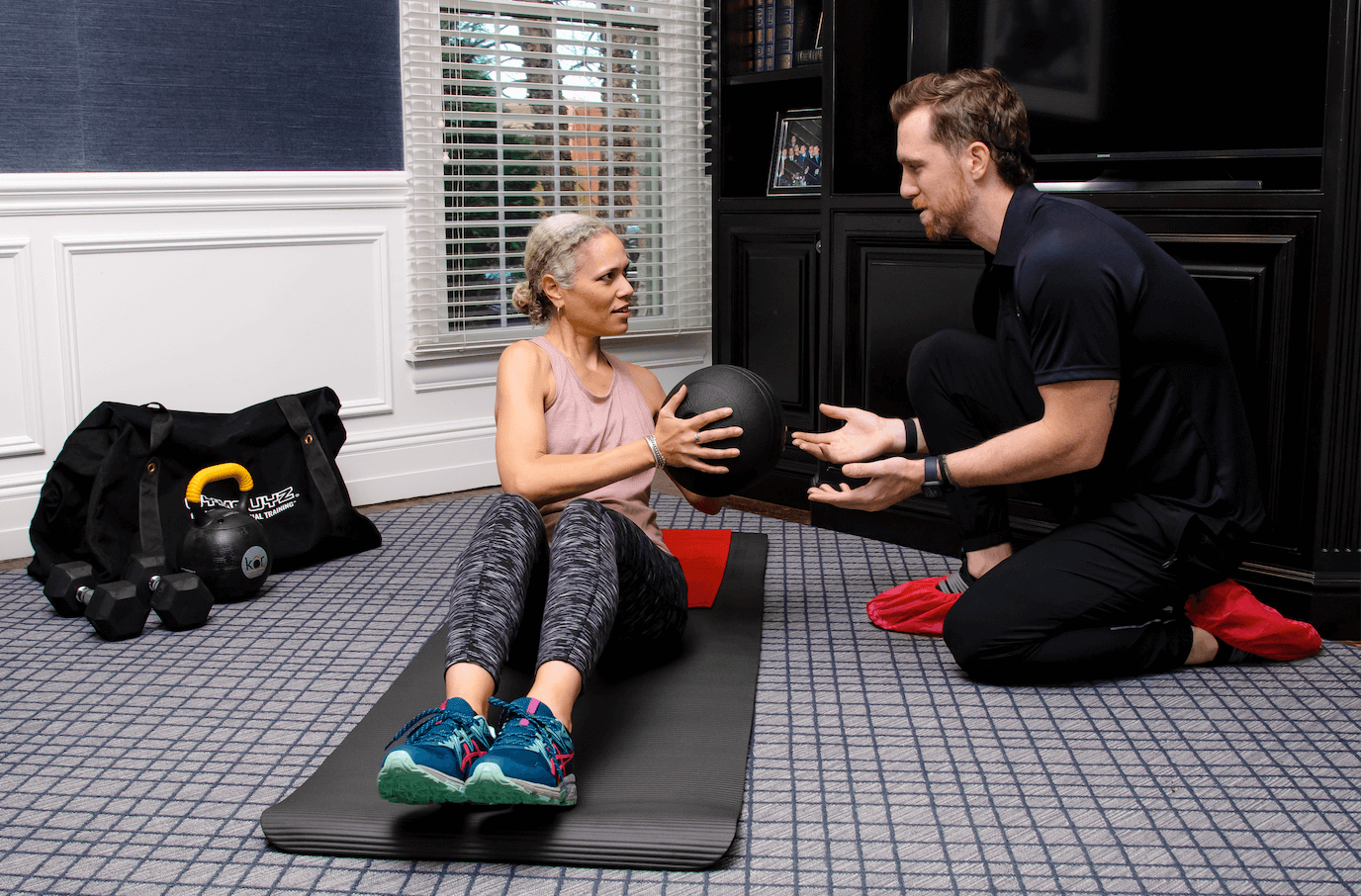 A woman is sitting on a yoga mat holding a medicine ball while a man kneels in front of her, explaining or instructing. They are in a room with a window, black cabinet, and fitness gear like dumbbells and a bag.