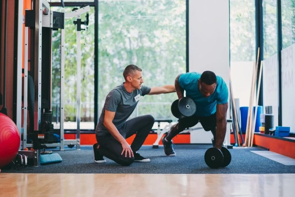 Personal trainer coaching a man doing a push-up with dumbbells in a gym.