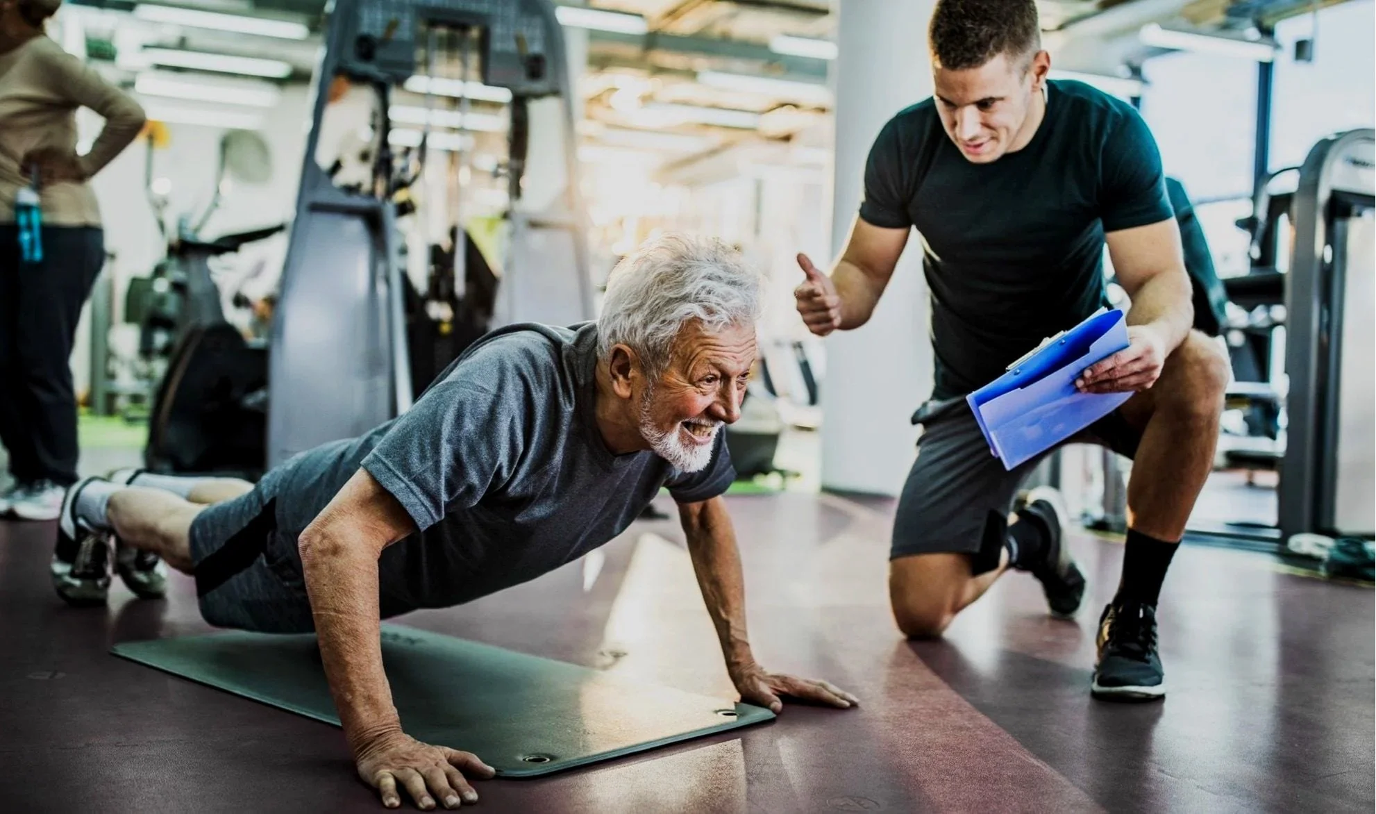 An older man doing a push-up on a mat at the gym with a trainer coaching him.
