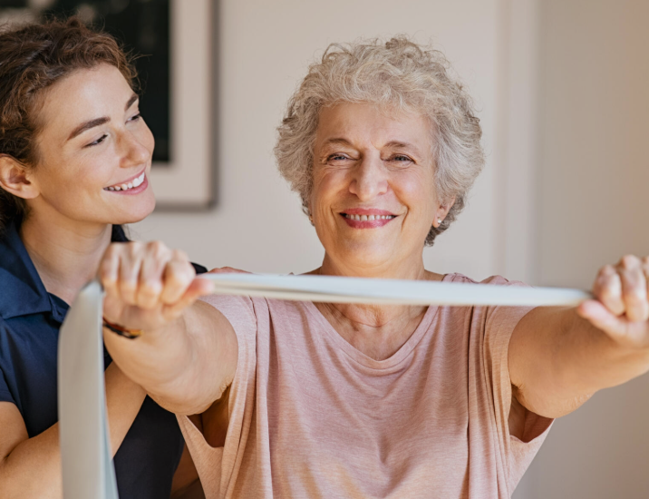 Older woman exercising with a resistance band while smiling, assisted by a younger woman.