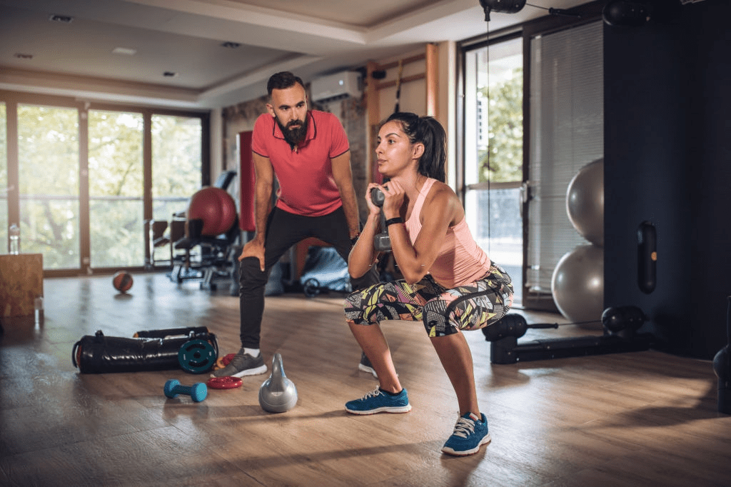 A woman squatting while holding a dumbbell in a gym, with a trainer observing her.