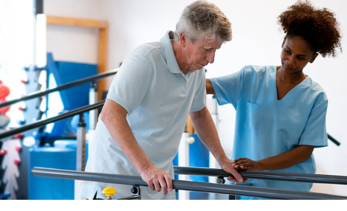 A man undergoing physical therapy with a therapist helping him walk with parallel bars.