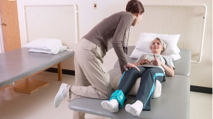 A healthcare worker assisting an elderly woman lying in a hospital bed, holding her hand and talking to her in a hospital room.