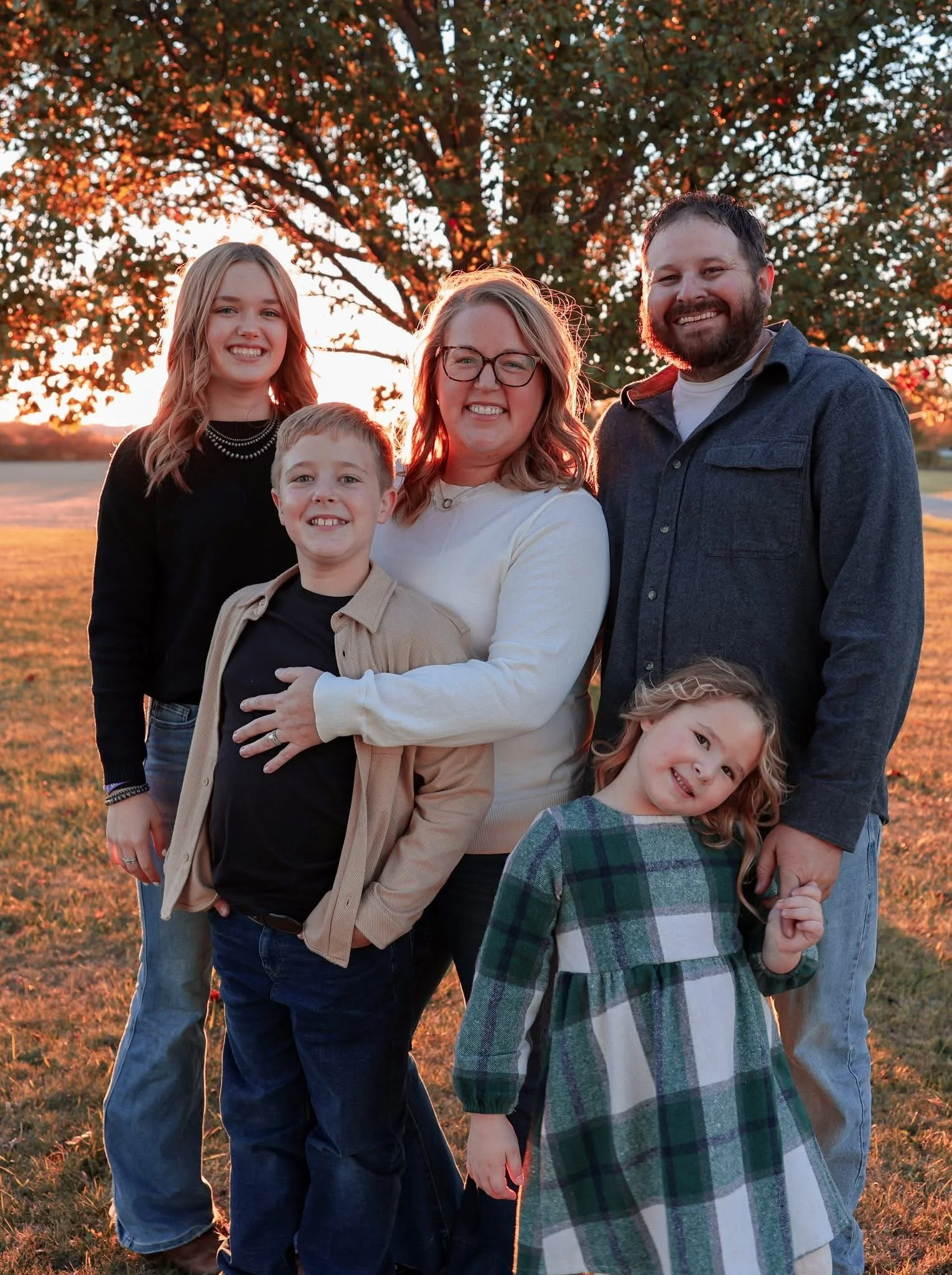 A family of six, including two women, two men, and two children, standing outdoors during sunset with a large tree and open field in the background. They are smiling and dressed casually.