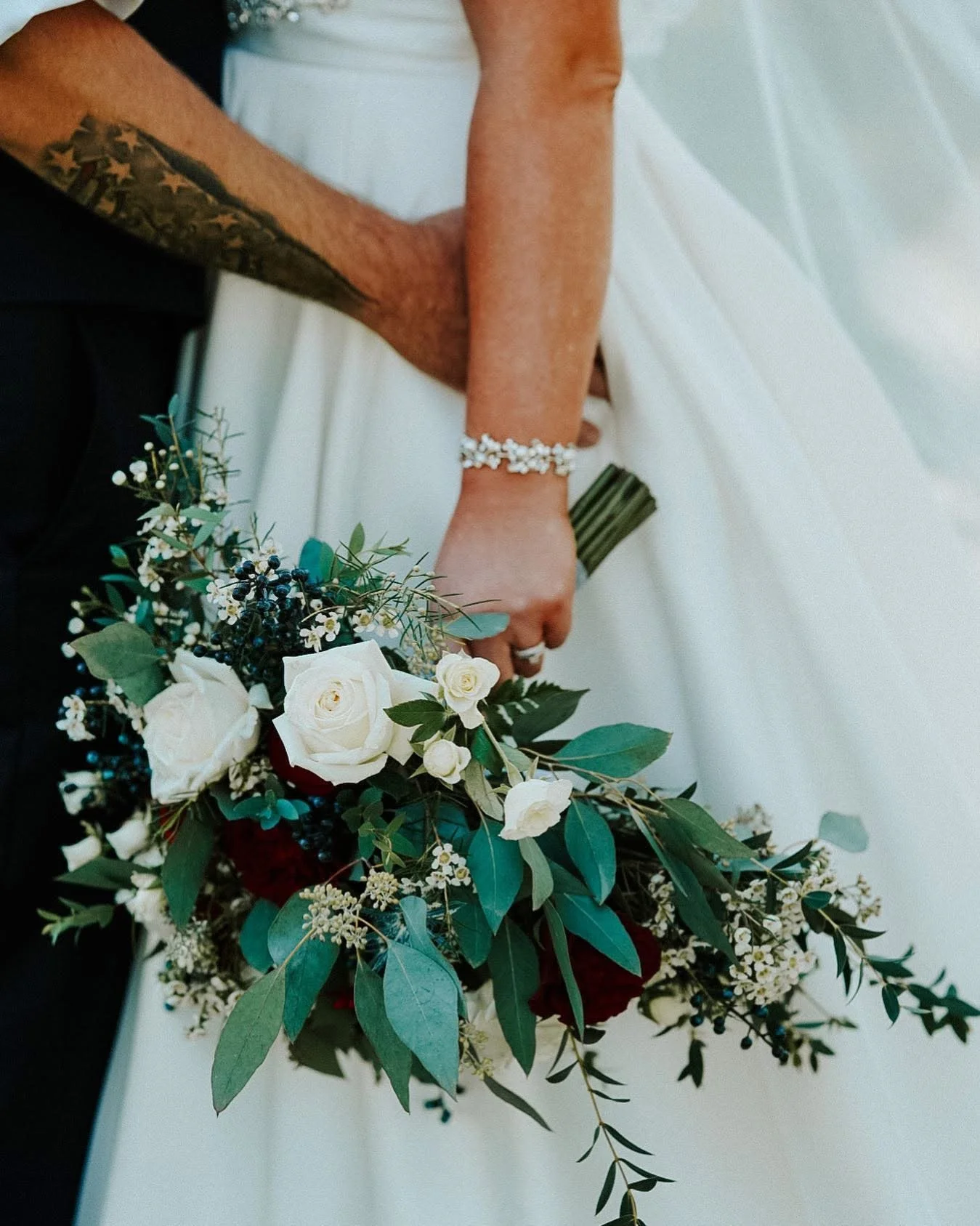 Close-up of a bride and groom holding hands, with the bride holding a bouquet of white and dark red roses and green foliage.