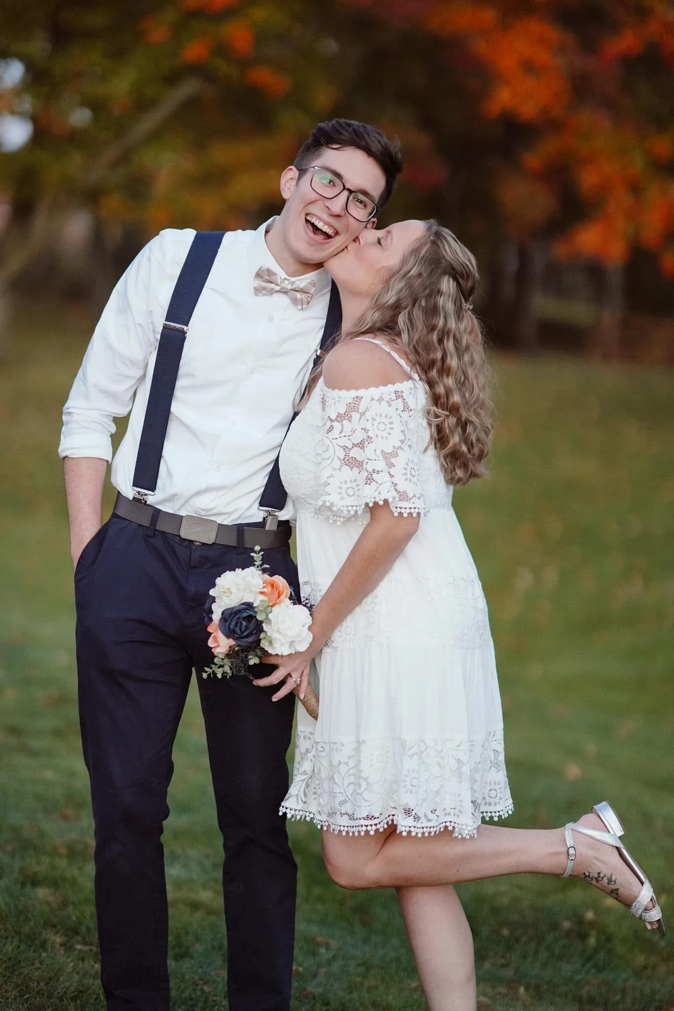 A couple in wedding attire outdoors, with the woman kissing the man's cheek while holding a bouquet, and the man smiling widely.