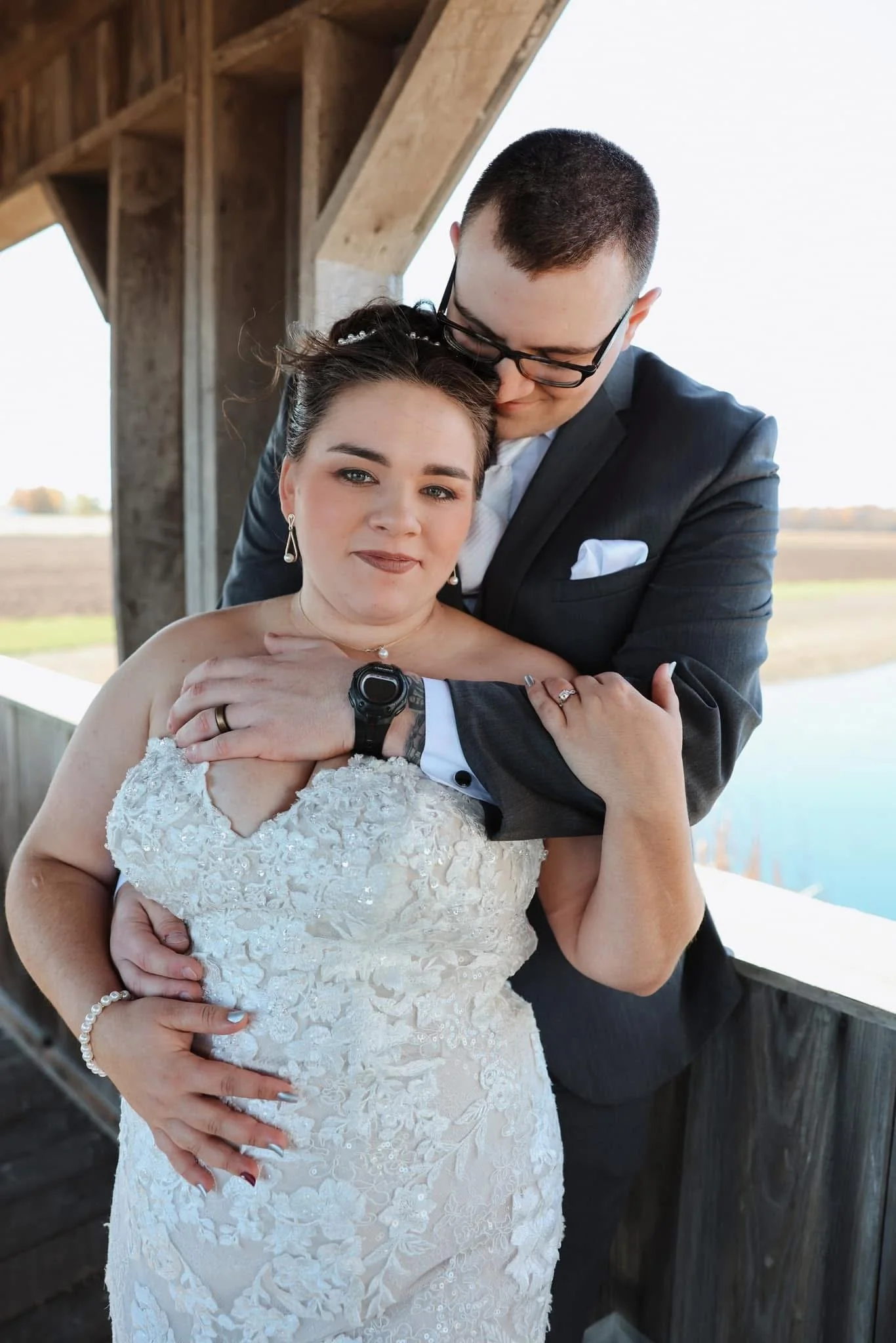 A bride and groom in wedding attire sharing an intimate moment outdoors with a wooden structure and open sky in the background.