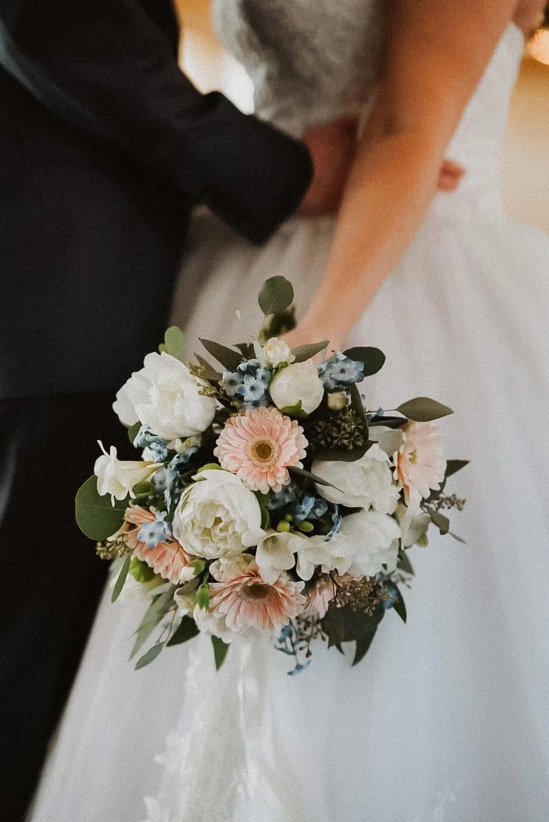 Close-up of a wedding bouquet with white, pink, and blue flowers being held by a bride in a white dress, with a groom in a black suit nearby.