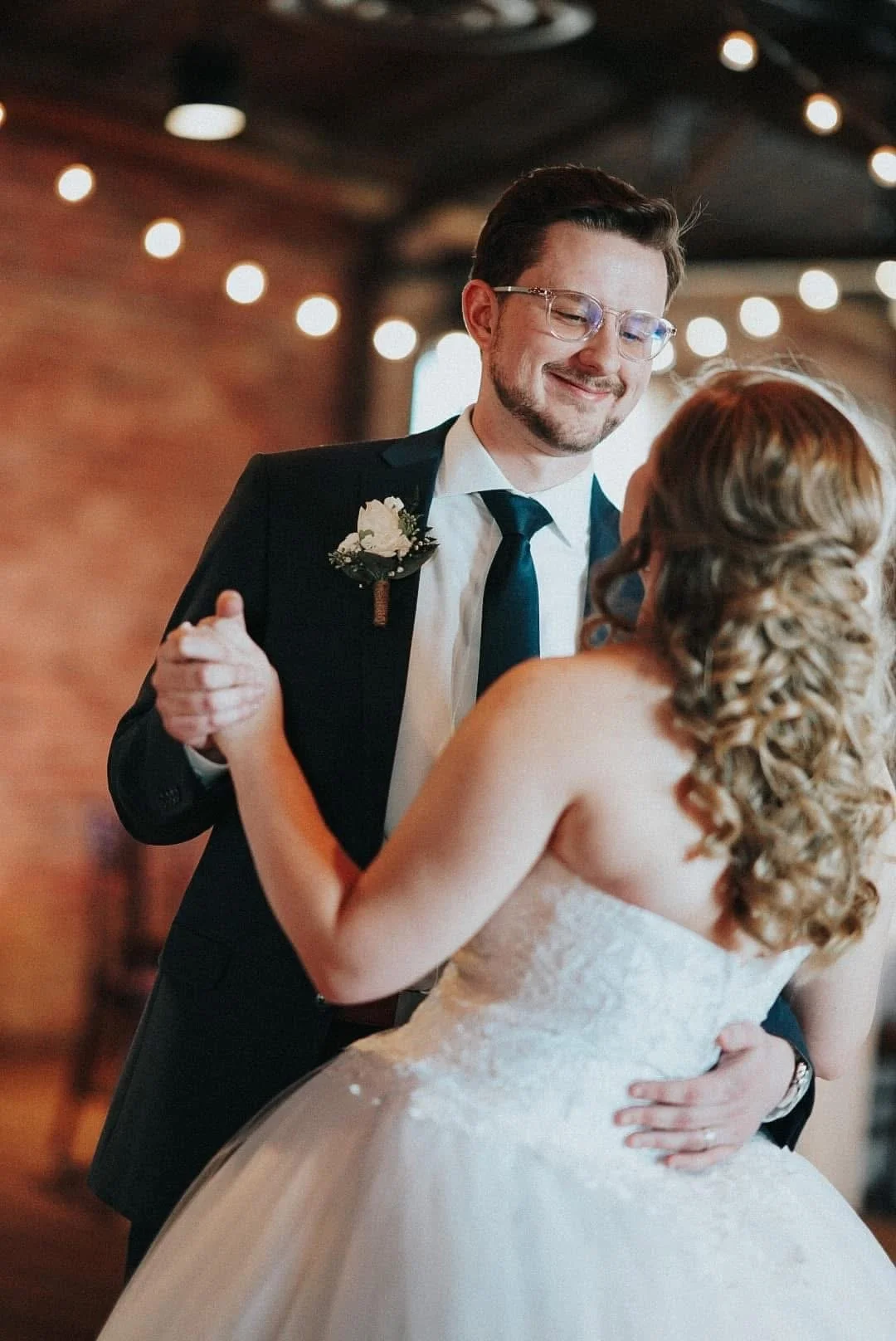 A groom in a suit and glasses dancing with a bride in a wedding dress, both smiling, during their wedding reception.