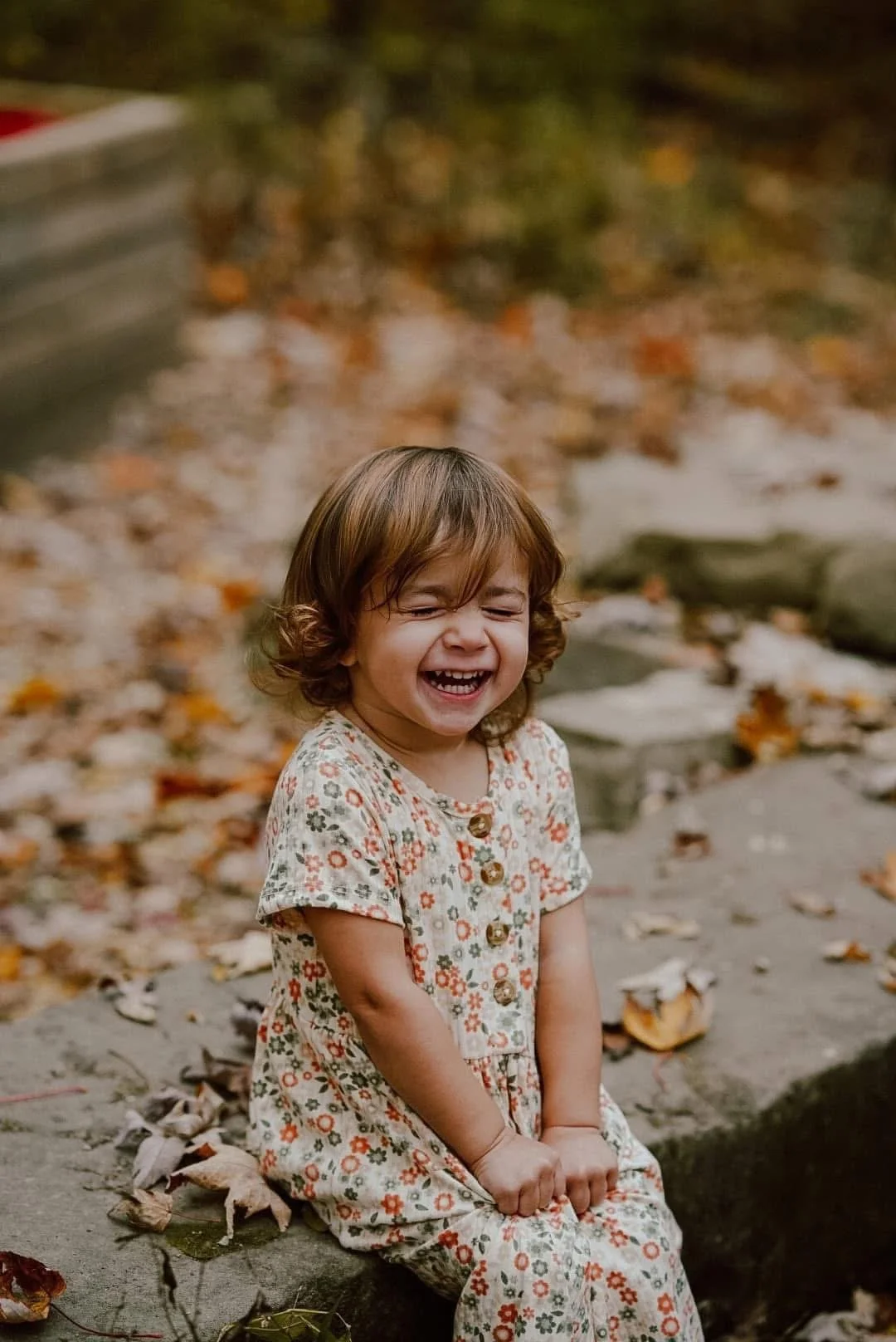 A young girl with brown, curly hair sitting outdoors on a rock, laughing with eyes closed, wearing a floral dress, surrounded by fallen autumn leaves.