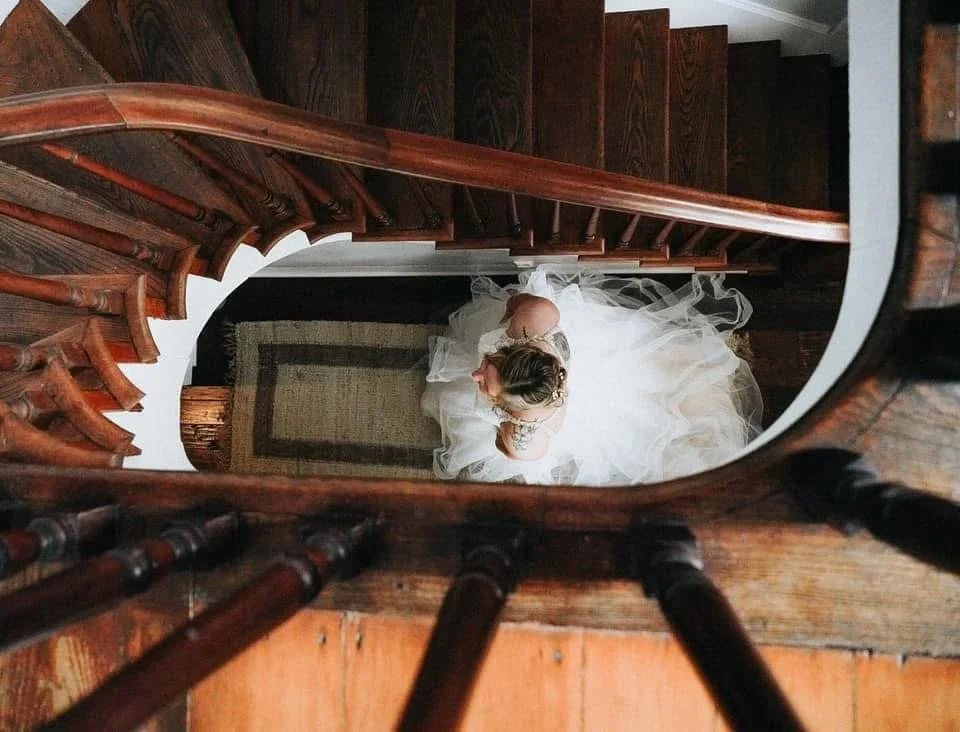 Top-down view of a bride in a wedding dress standing on a wooden staircase, captured through the banister.