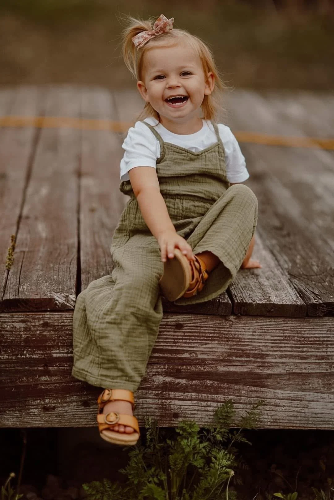 A young girl with blonde hair, smiling and sitting on a wooden dock, wearing a white T-shirt, olive-green overalls, and yellow sandals, with a pink bow in her hair.