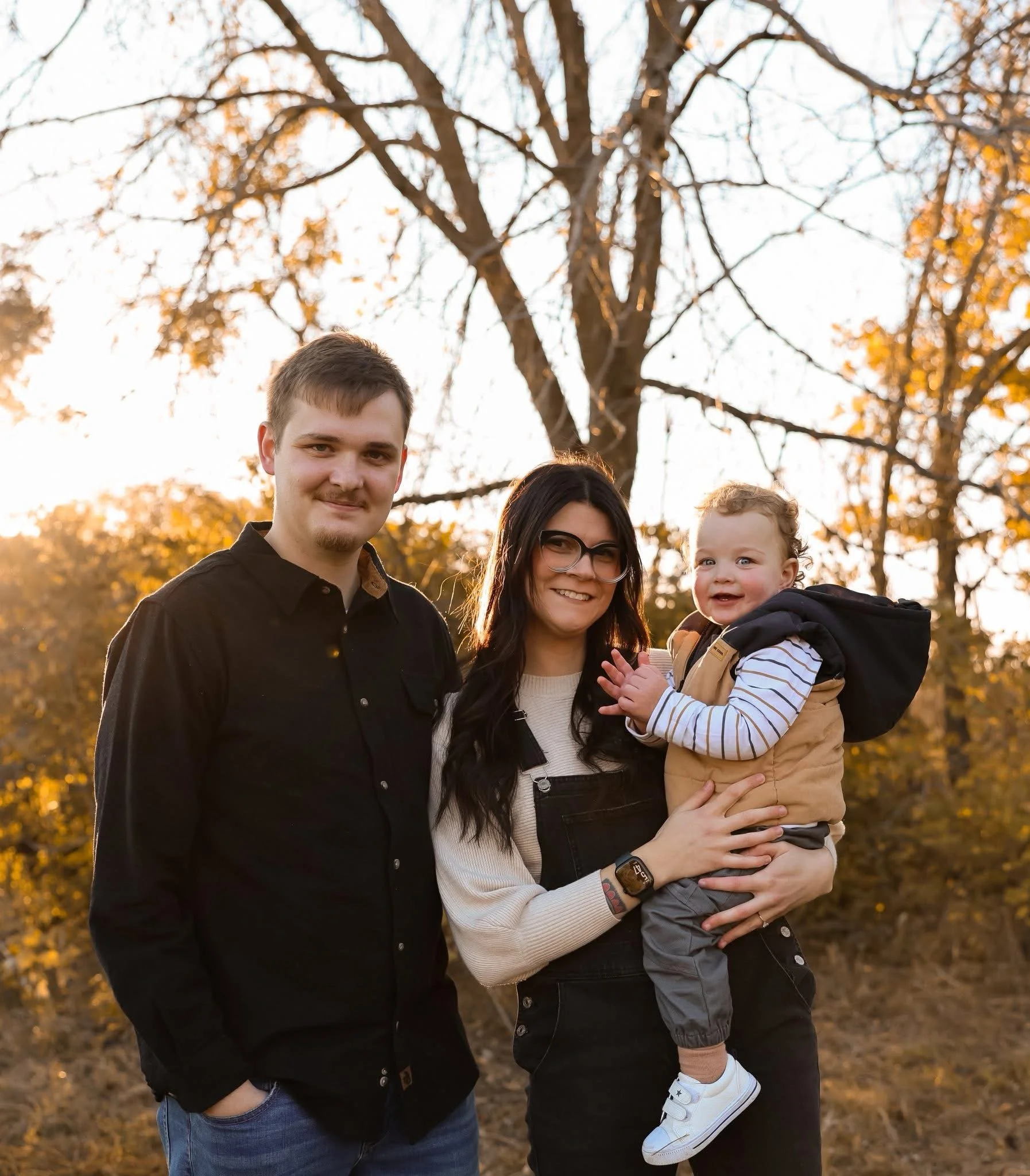 A family of three standing outdoors during autumn, with trees and fall foliage in the background. The woman holds a young child, and the man stands next to them smiling.