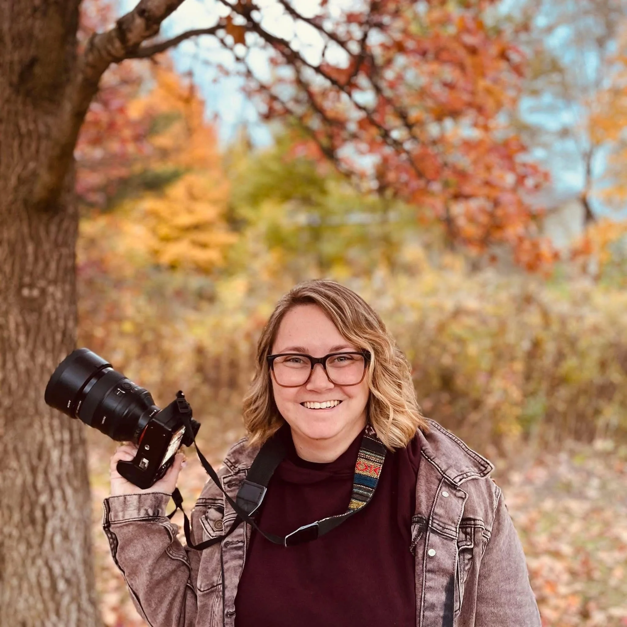 A smiling woman with glasses holding a camera on her shoulder outdoors in autumn, with colorful fall leaves in the background.