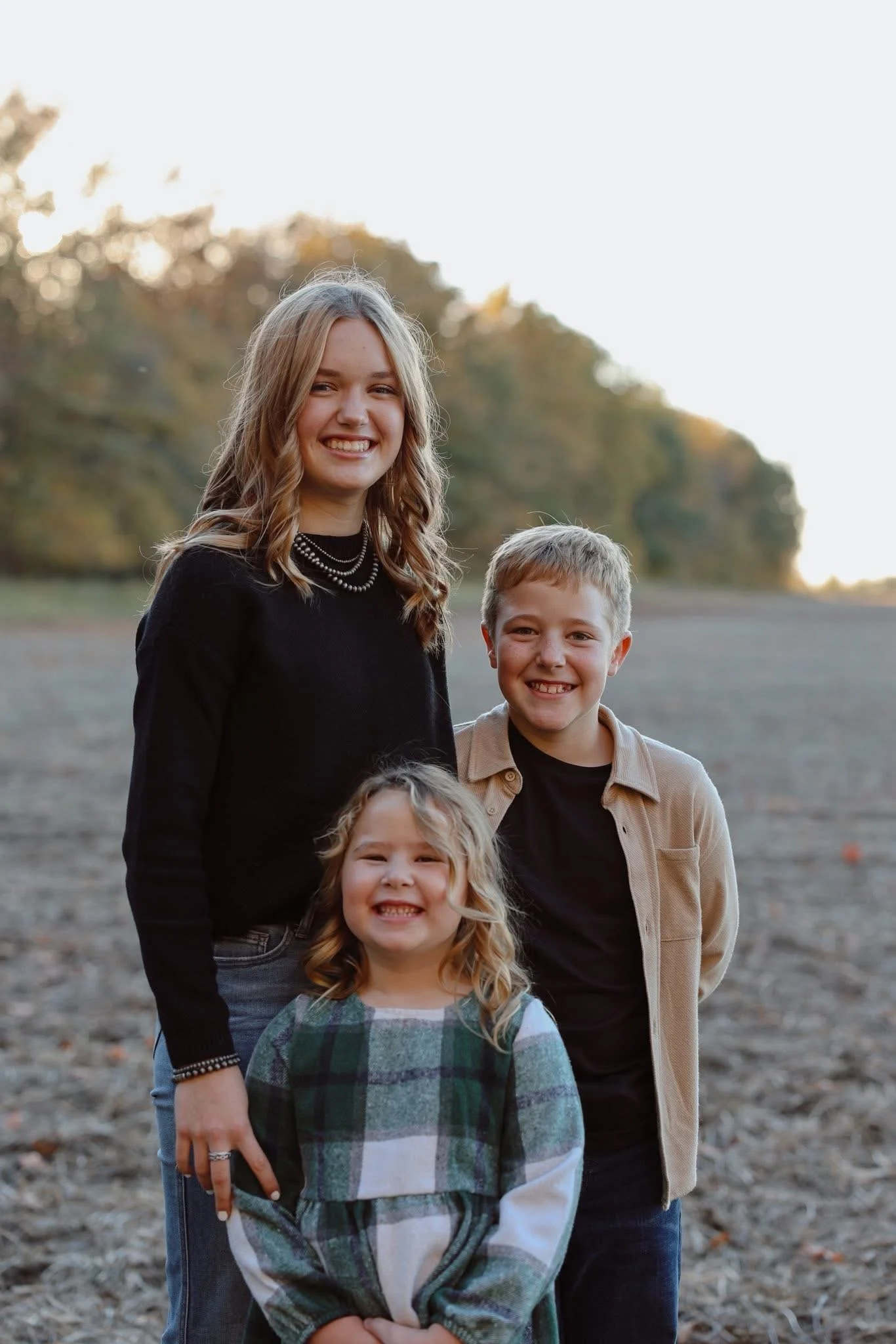 A group of three children smiling outdoors in a field with a background of trees during sunset.