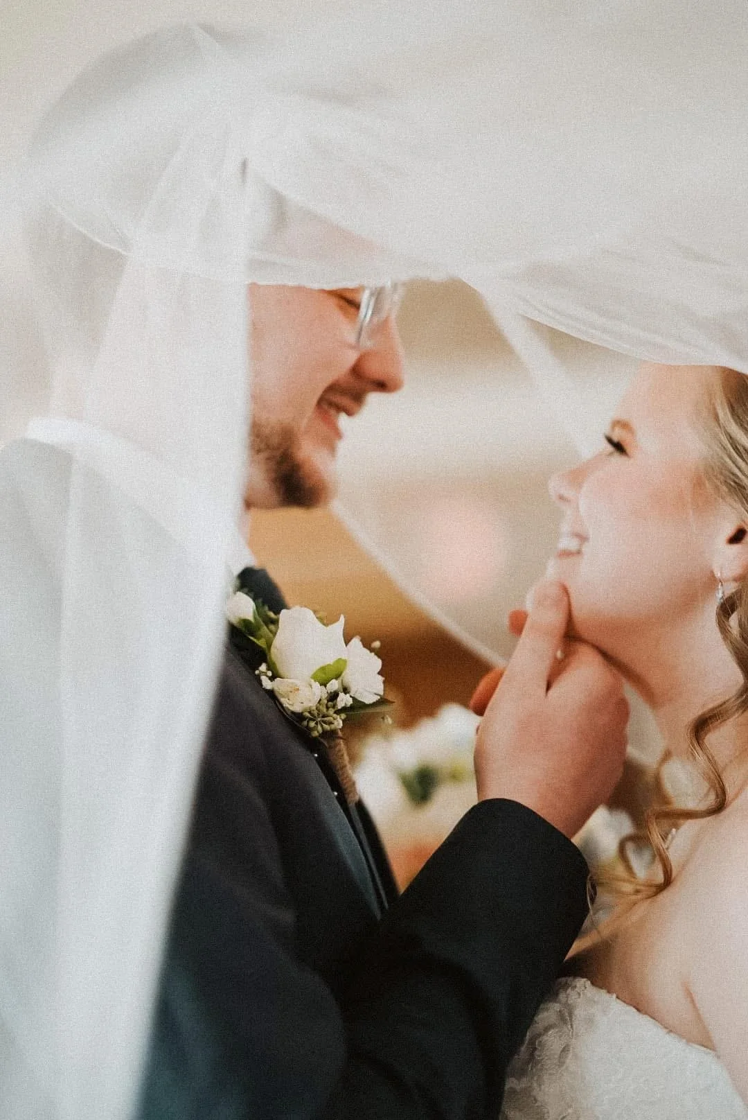 A bride and groom share a joyful moment under the wedding veil, with the groom gently touching the bride's chin as they smile at each other.