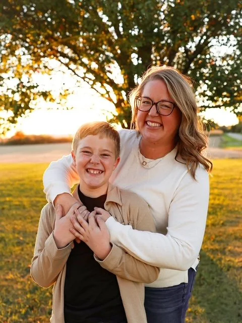 A woman with glasses and a young boy smiling and hugging outdoors during sunset, with a tree and open field in the background.