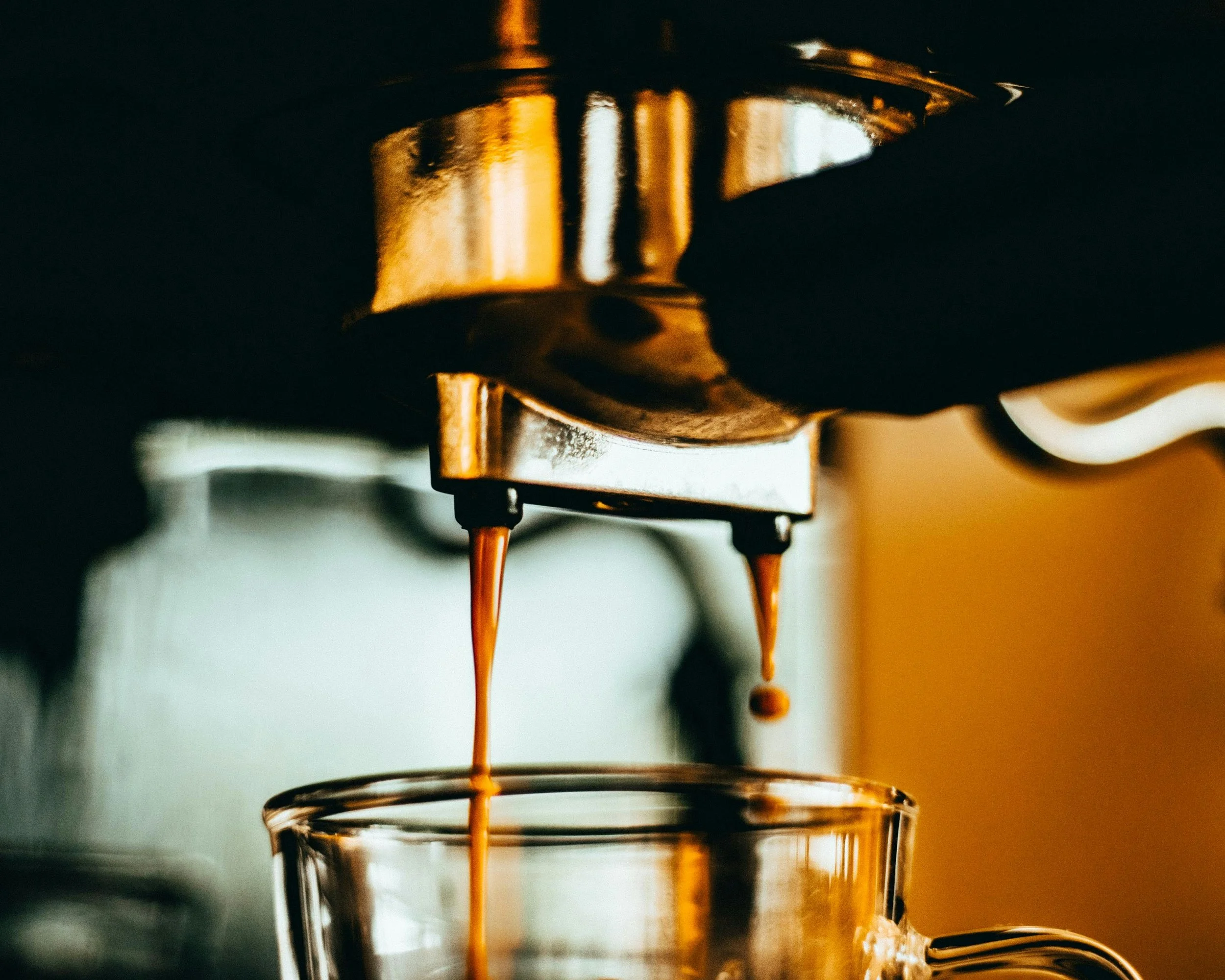 Close-up of espresso being brewed, with hot coffee dripping from the espresso machine spout into a clear glass cup.