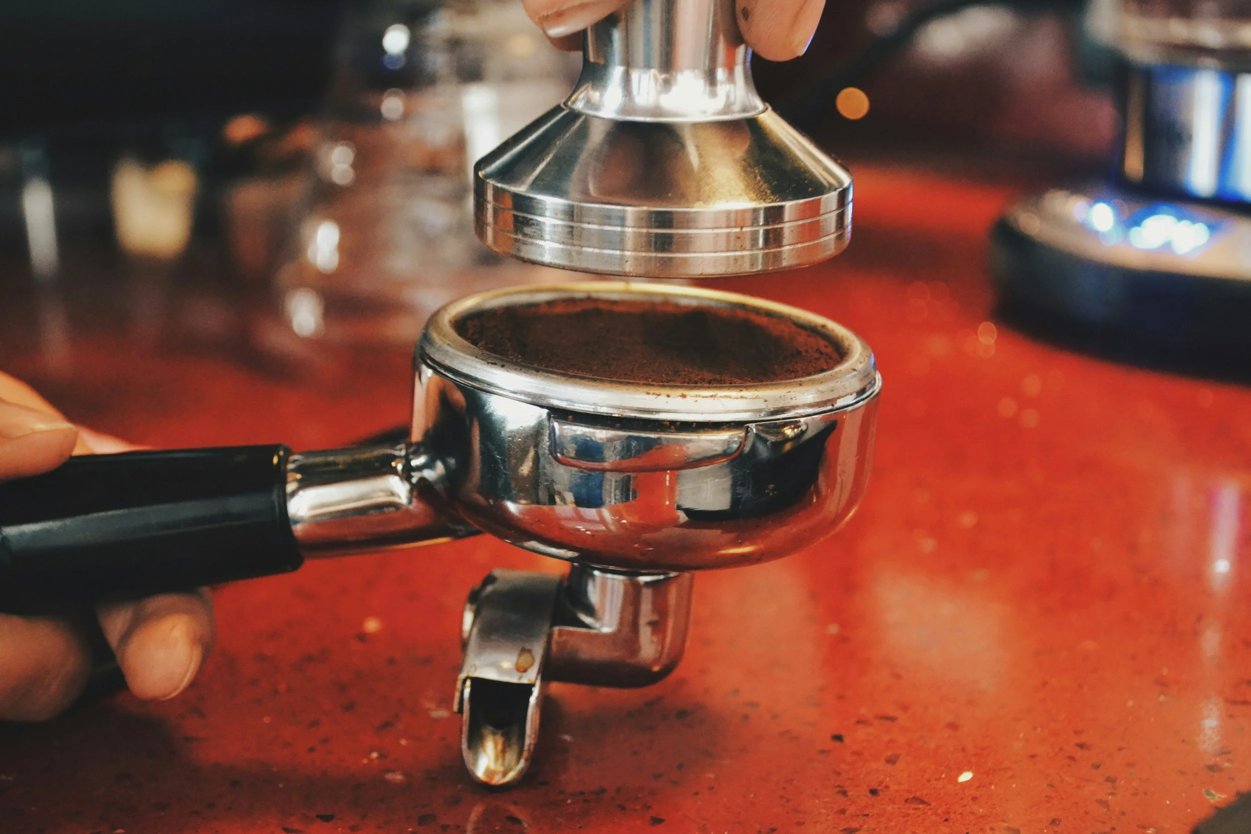 Close-up of a coffee tamper pressing coffee grounds in a portafilter on an orange countertop, with a metal distribution tool above.
