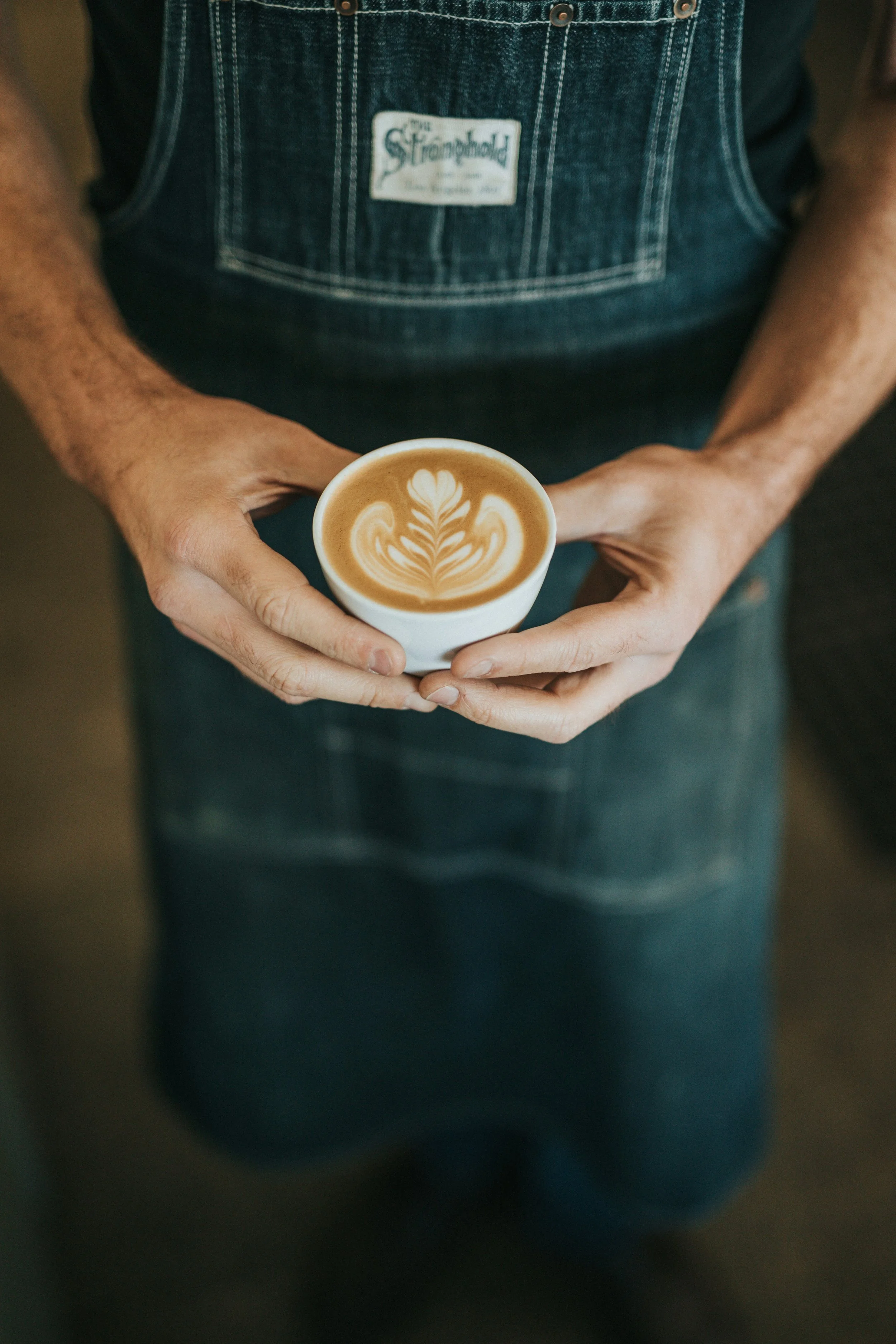 Person holding a cup of latte with latte art on top, wearing a denim apron.
